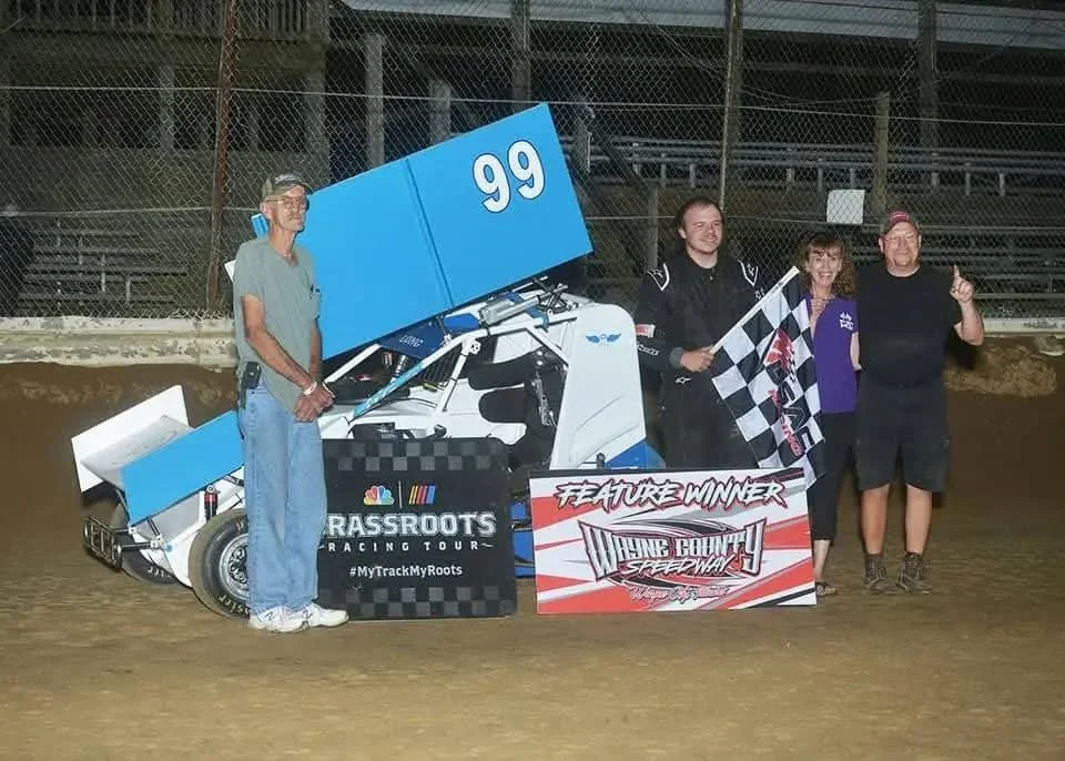 Group of four people celebrating a victory at a dirt race track, with a race car, checkered flag, and a sign that reads "Feature Winner" and "June Bounty Speedway"