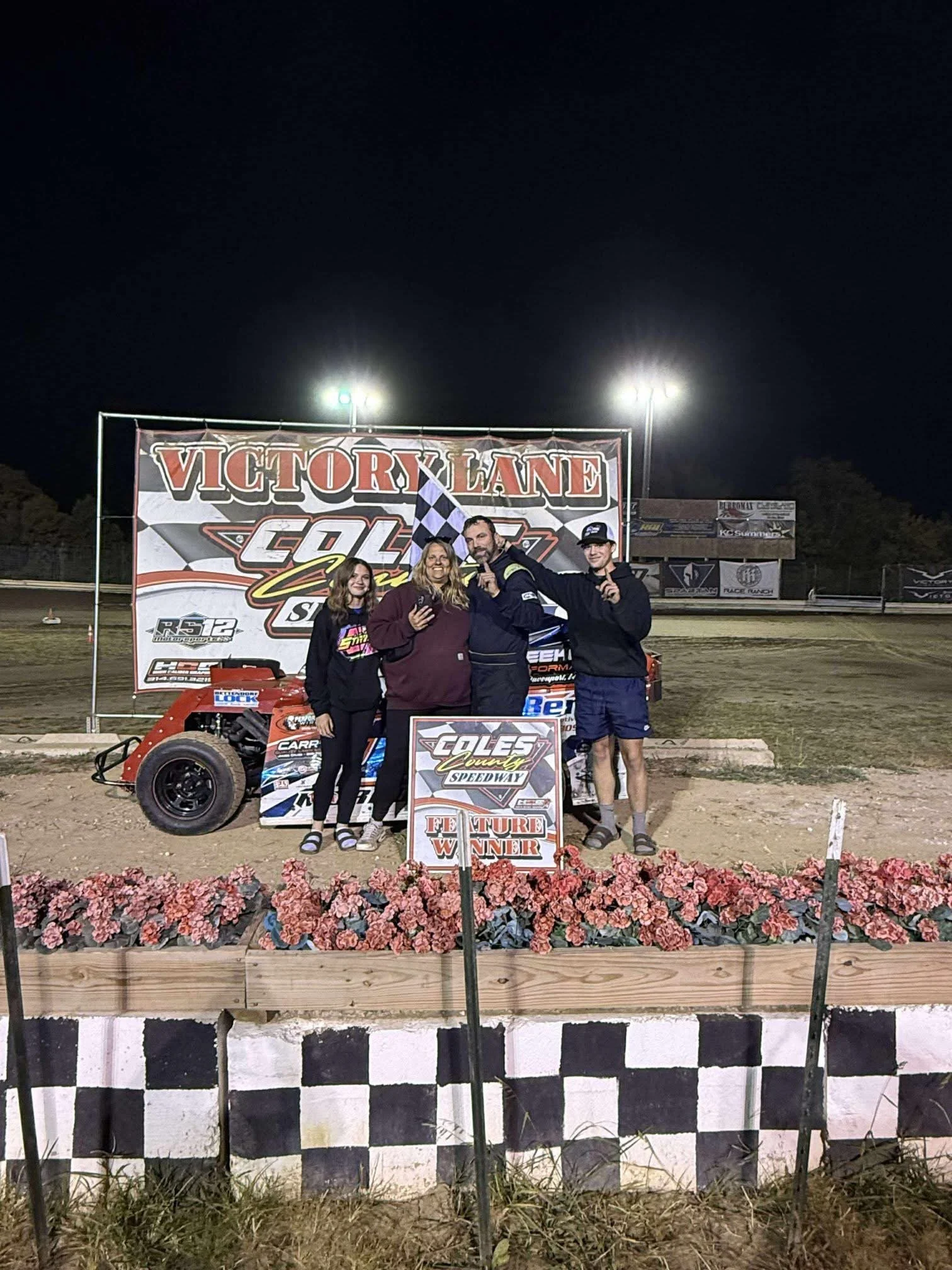 Four people celebrating a racing victory at the Cole County Speedway, with a small race car and a large victory banner in the background.