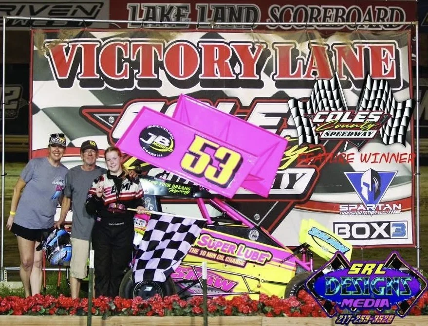 A girl in racing gear standing next to a pink modified race car with the number 53, holding a checkered flag at Victory Lane after a race at Lake Land Speedway. Three people are standing behind her, and a large banner with various sponsor logos and t