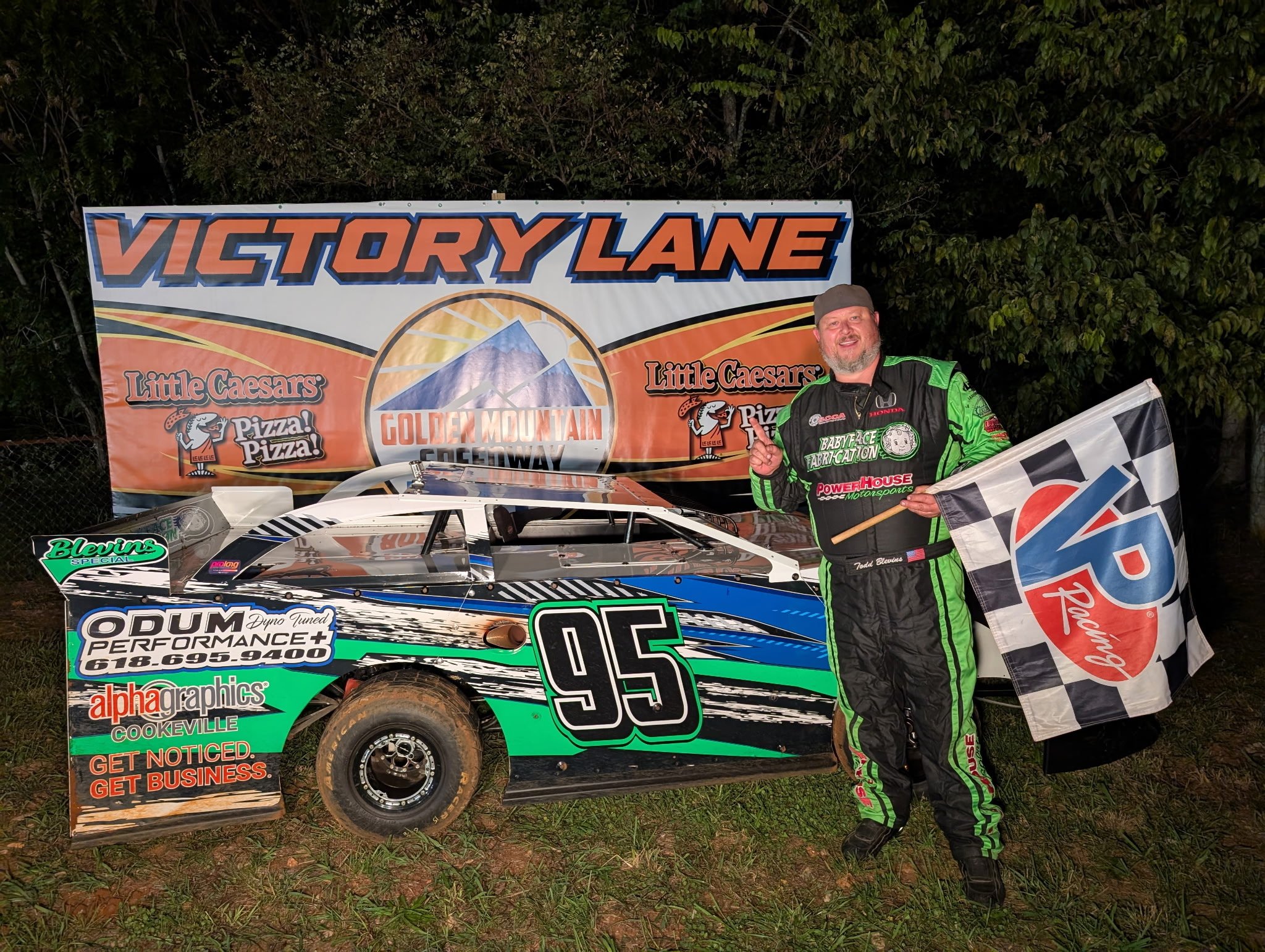 Race car driver standing next to a race car holding a checkered flag, at Victory Lane celebrating a win, with a large banner in the background displaying course and sponsors.