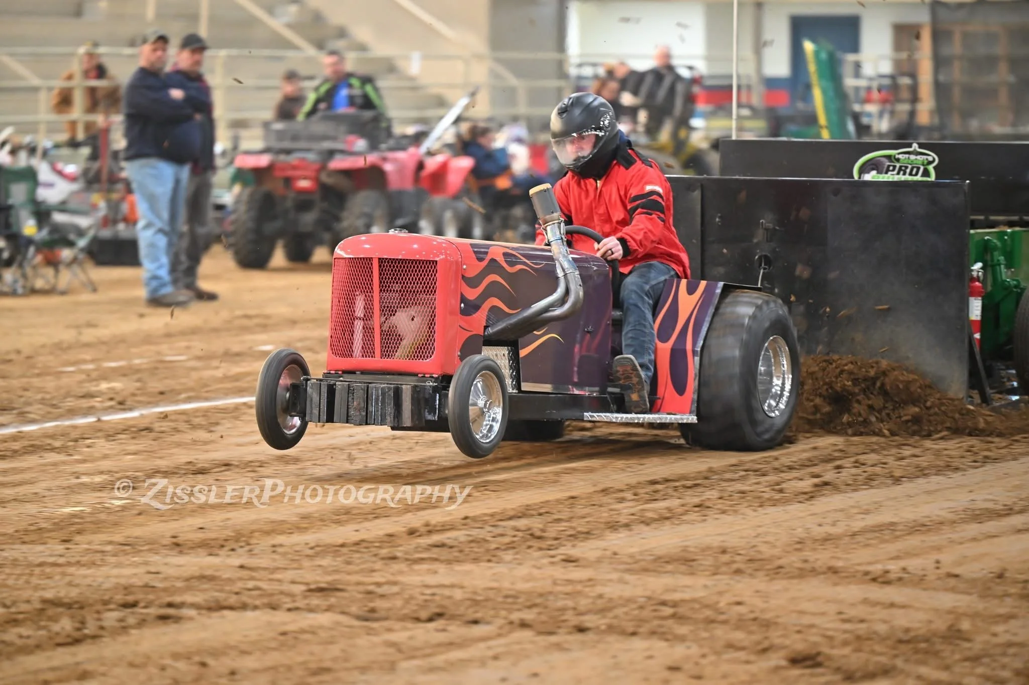 A young boy in a red jacket and black helmet driving a custom mini tractor racing on a dirt track during an event, with spectators and vehicles in the background.