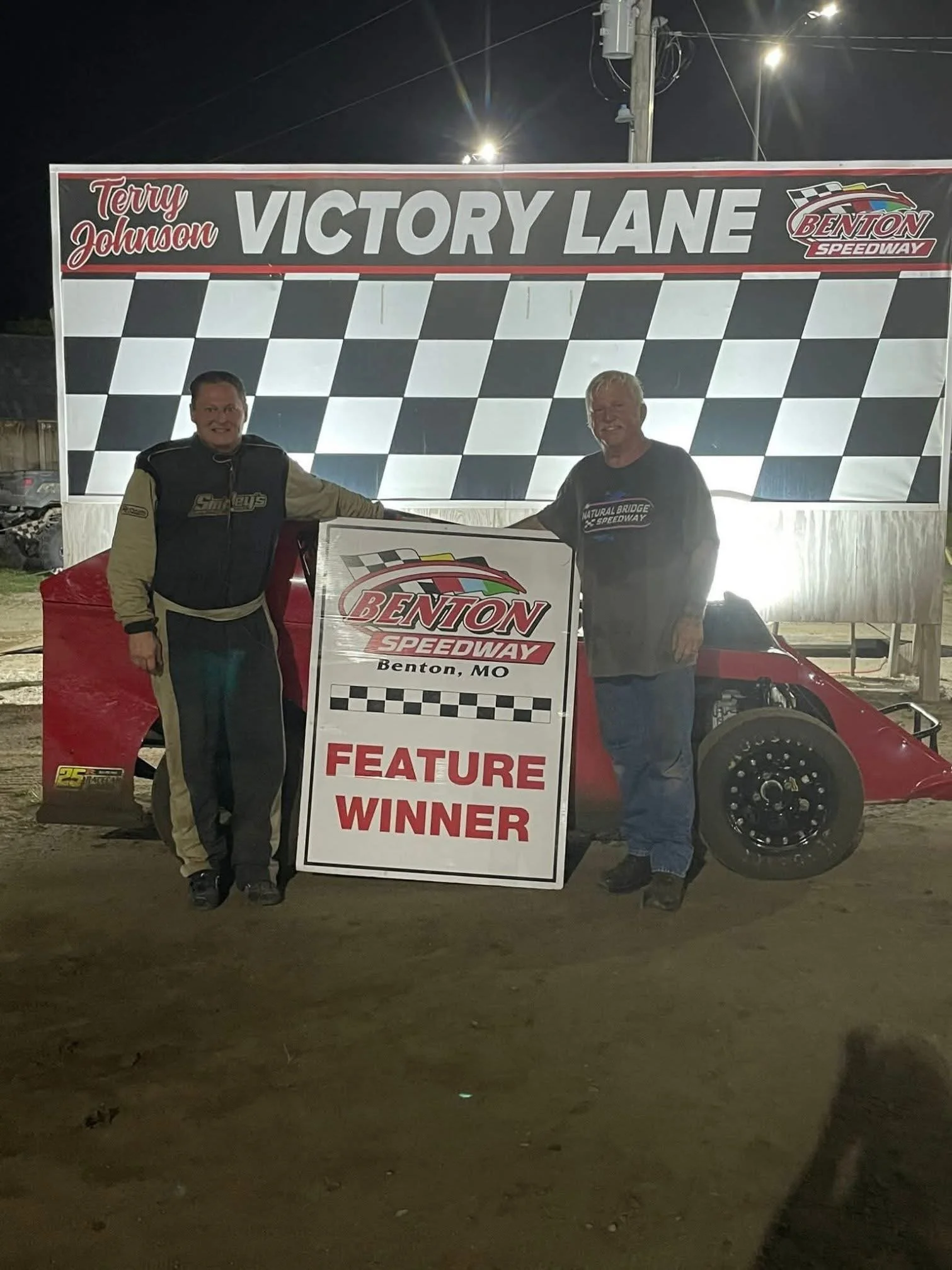Two men standing in front of a race car at Benton Speedway in Benton, Missouri, holding a sign that says "Feature Winner." The backdrop reads "Victory Lane" with checkered flags and the name "Terry Johnson." The event appears to be a nighttime race c