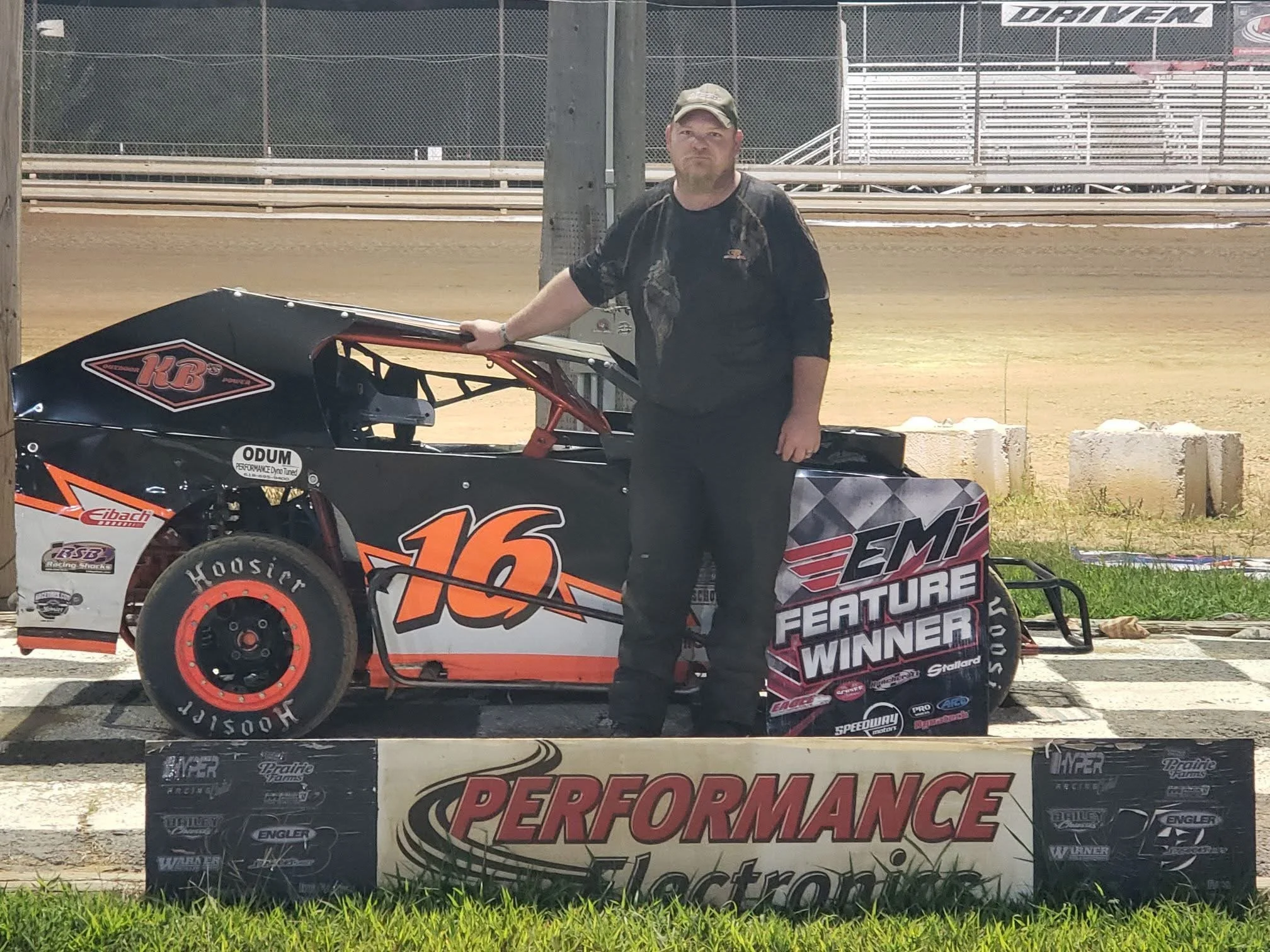 A man standing next to a black and orange race car with the number 16, holding a sign that says 'EMI FEATURE WINNER,' on a racetrack.