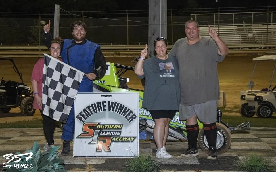 Four people standing together at a racing track with a sprint car, celebrating a win. The person in the blue racing suit holds a checkered flag and a sign that says 'Feature Winner Southern Illinois Raceway.' Two people are holding up their index fin