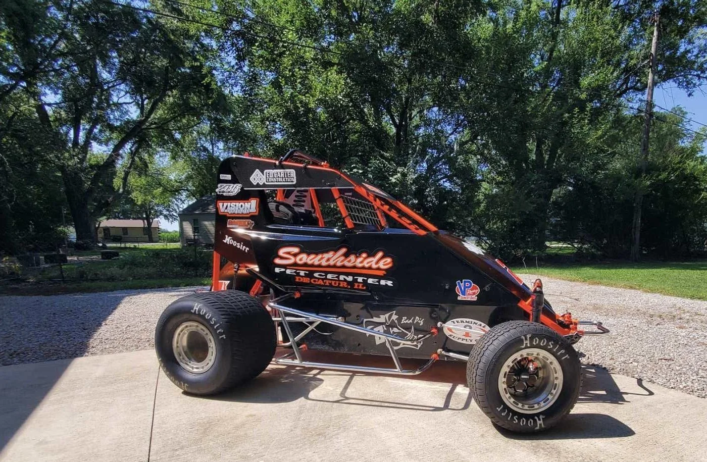 A black and orange quarter midget race car with sponsors and decals, parked on a concrete driveway with trees and a house in the background.