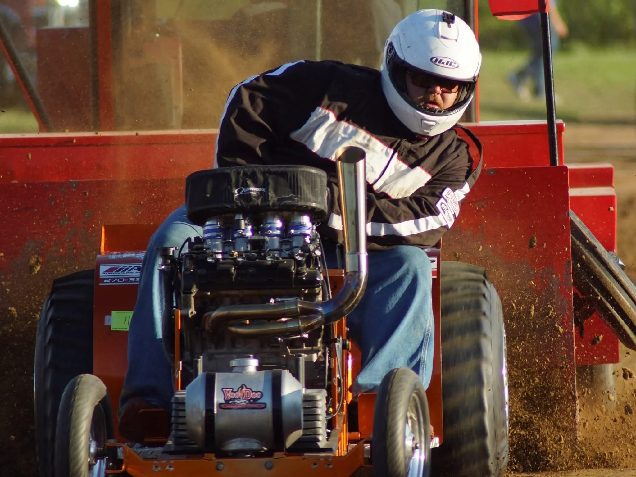 A person wearing a white helmet, sunglasses, and racing jacket driving a lawnmower on a dirt track.