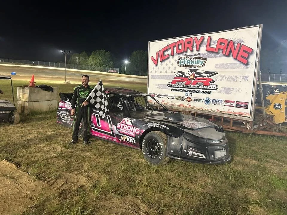 Race car driver standing next to a black and pink race car holding a checkered flag at Victory Lane after a race, at a dirt race track at night.