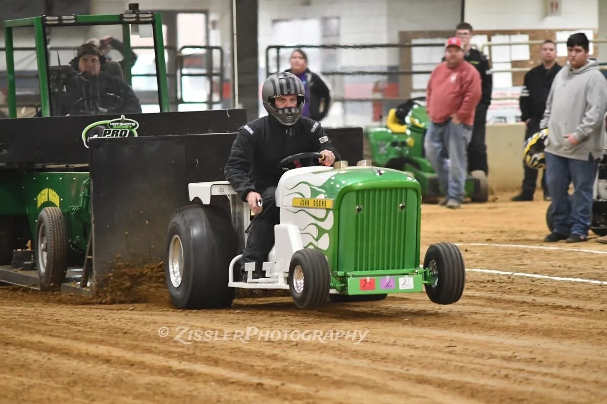 A person driving a miniature tractor with a green and white color scheme on an indoor dirt track, with several onlookers watching in the background.
