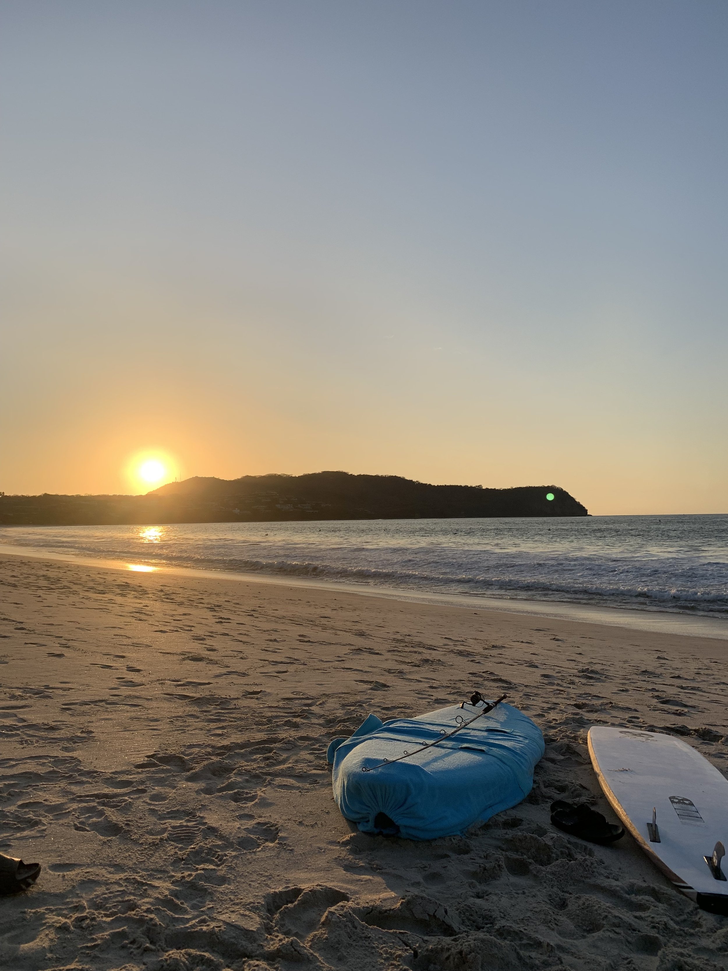 Beach scene at sunset with a packed blue beach bag and a paddleboard lying on the sand.
