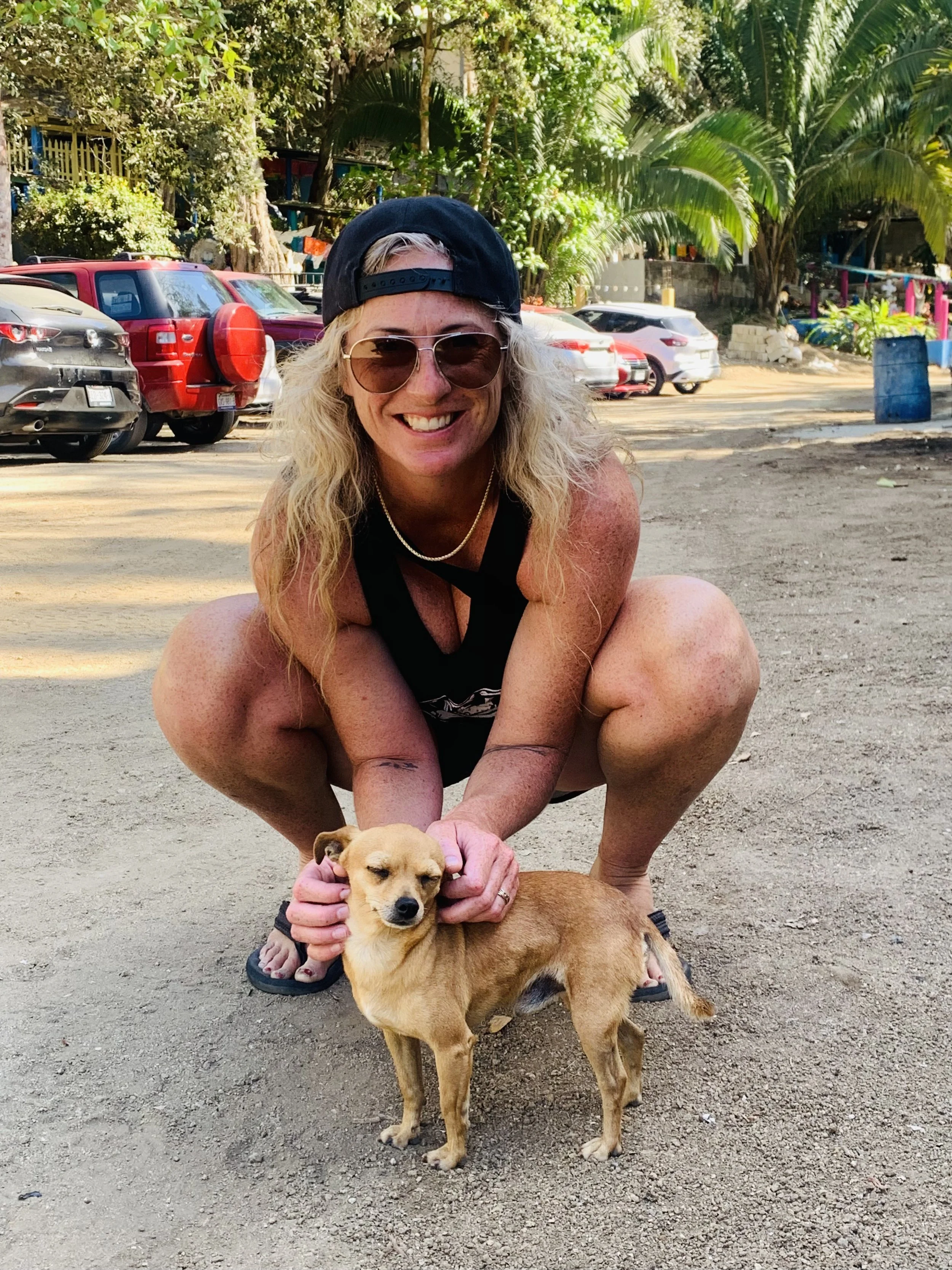A woman squatting and smiling while holding a small tan dog outdoors in a parking lot with trees and several parked cars in the background.