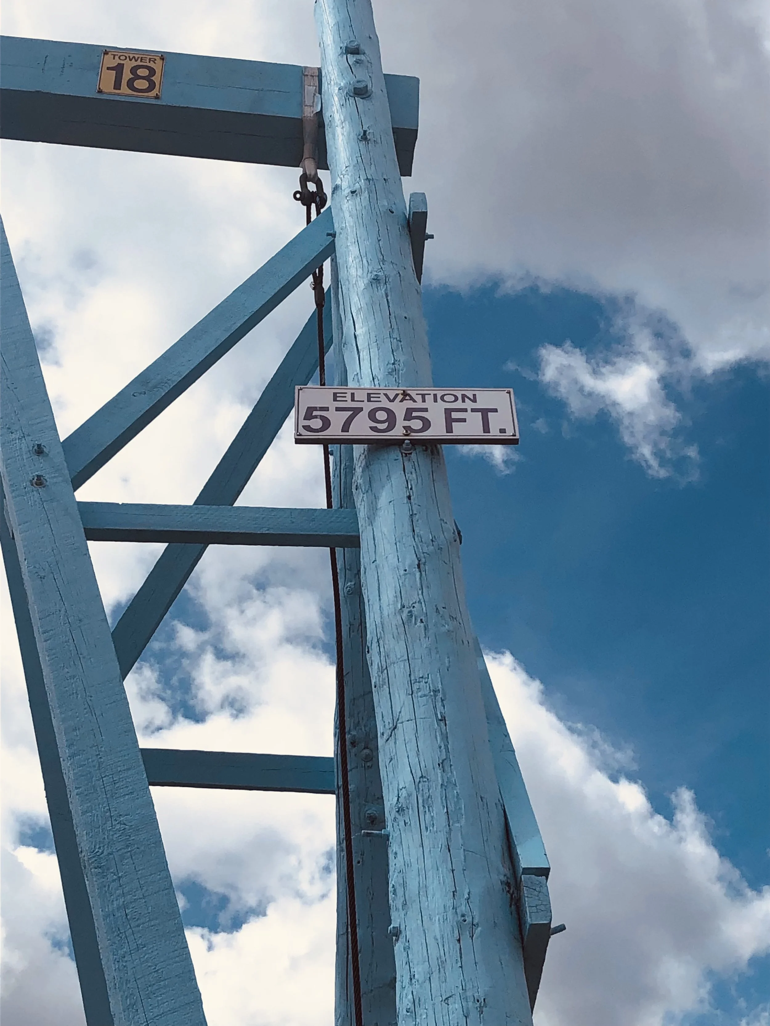 A tall wooden utility pole painted blue with a sign showing an elevation of 5795 feet, and part of a blue-painted structure marked 'Tower 18' at the top, against a partly cloudy sky.