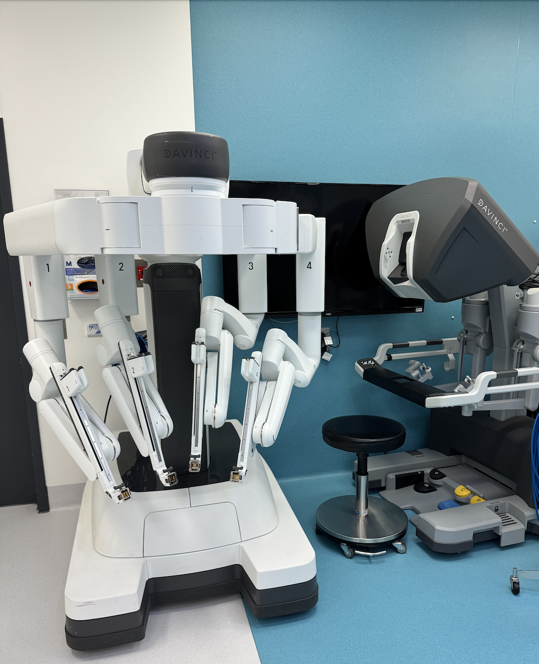 Medical robotic surgical system with multiple robotic arms and a large screen, located in a clinical room.