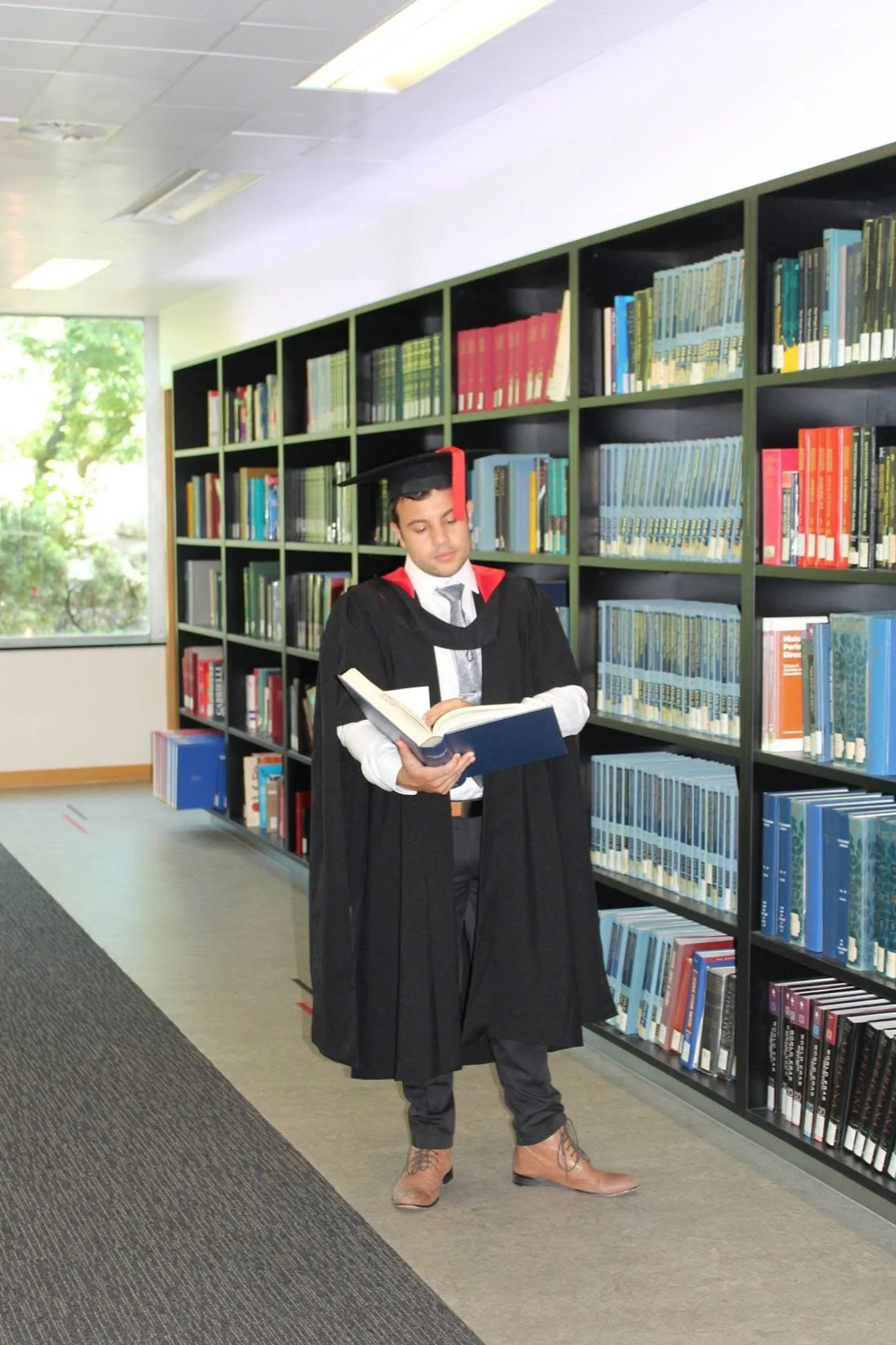 Young man in a graduation gown and cap reading a book in a library.