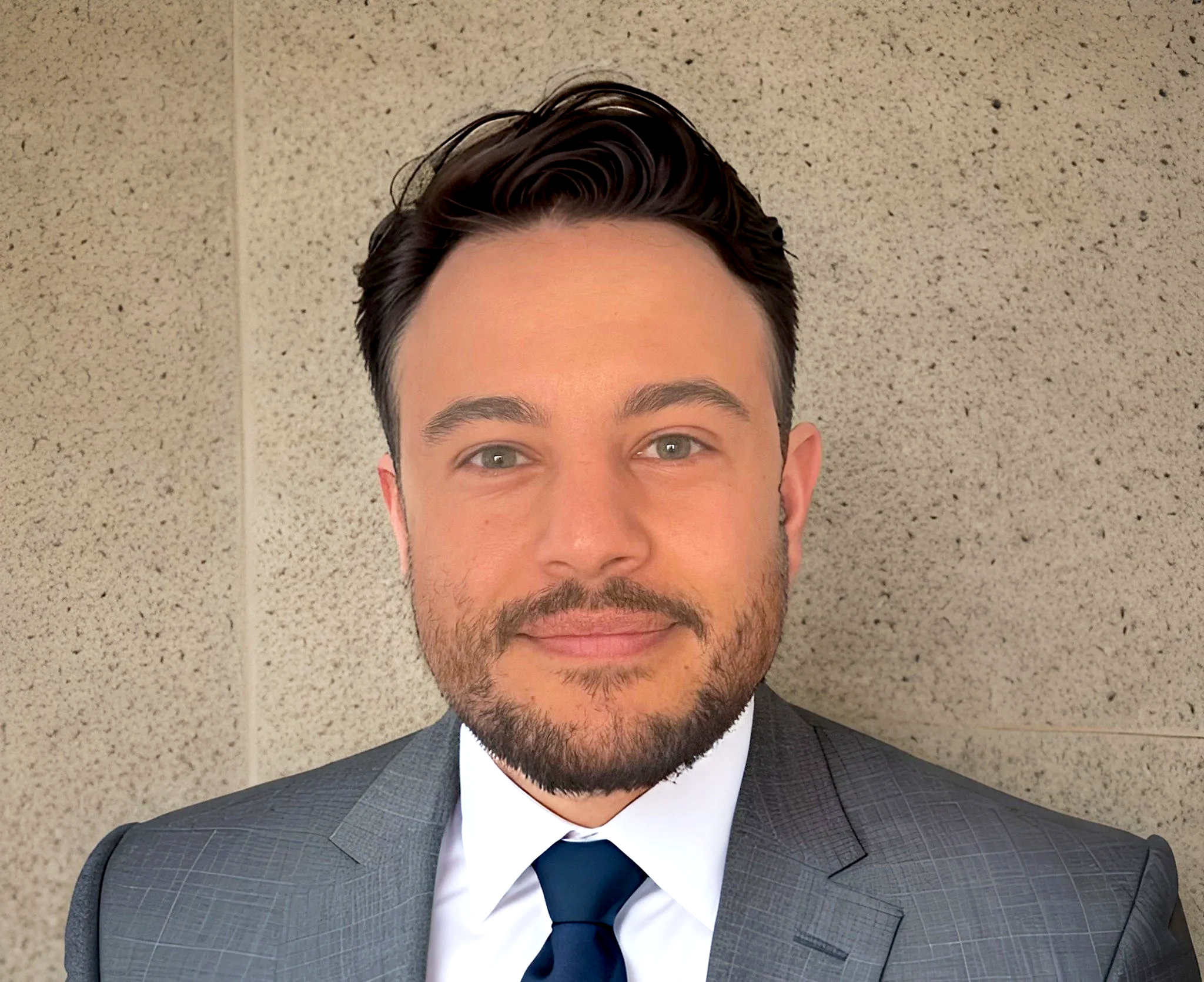 A man with dark hair, a beard, and light skin, wearing a gray suit, white shirt, and dark tie, smiling in front of a textured beige wall.
