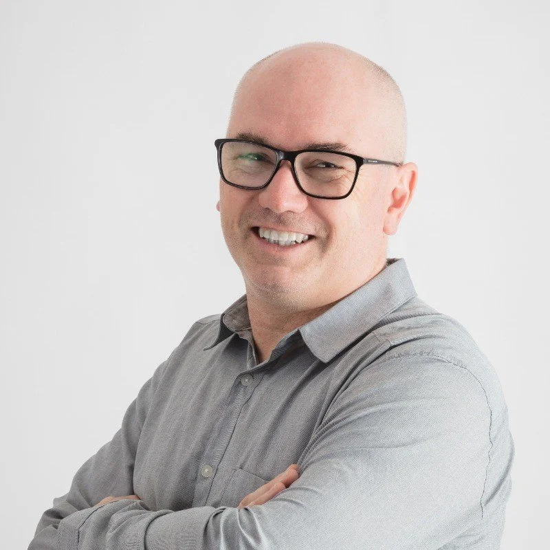 Headshot of a bald man wearing glasses and a gray button-up shirt, smiling with arms crossed against a plain white background.