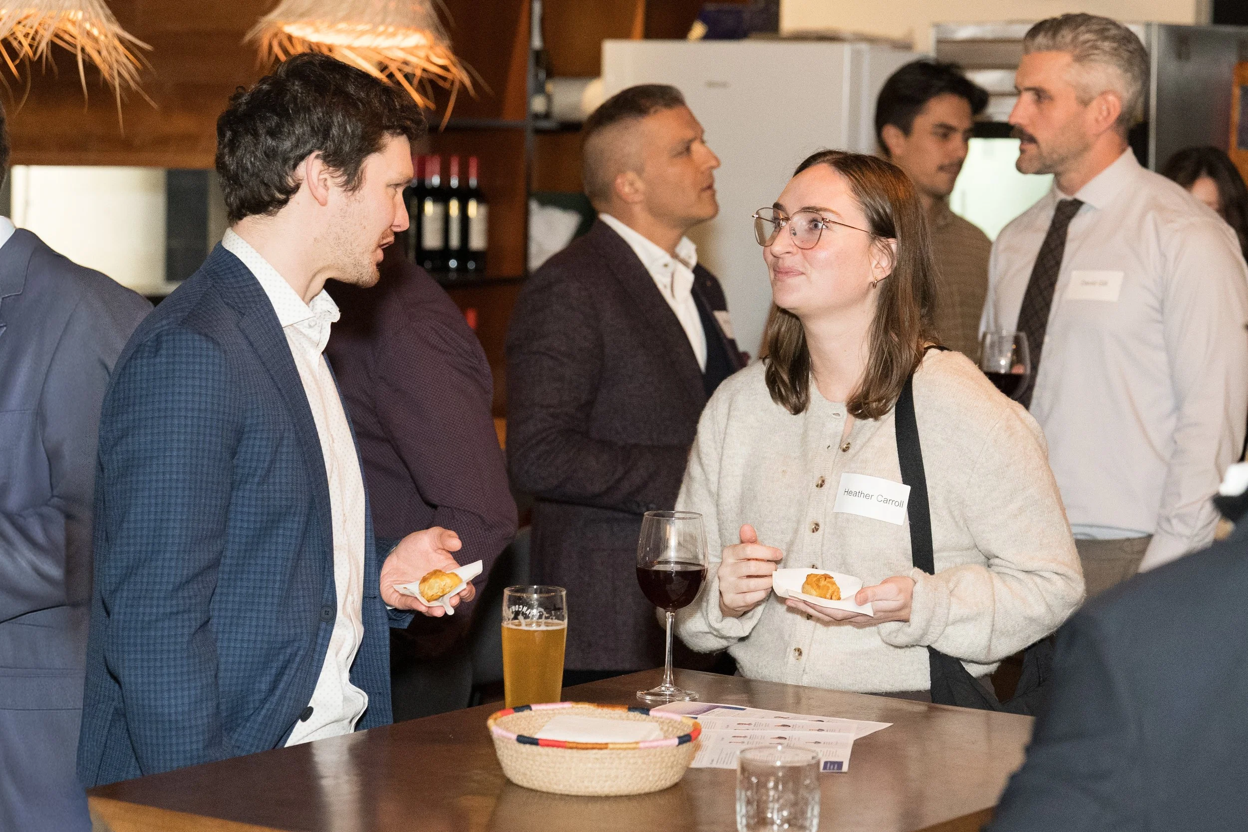 People socializing at a gathering, standing around a table with food and drinks in a cozy, wooden interior.