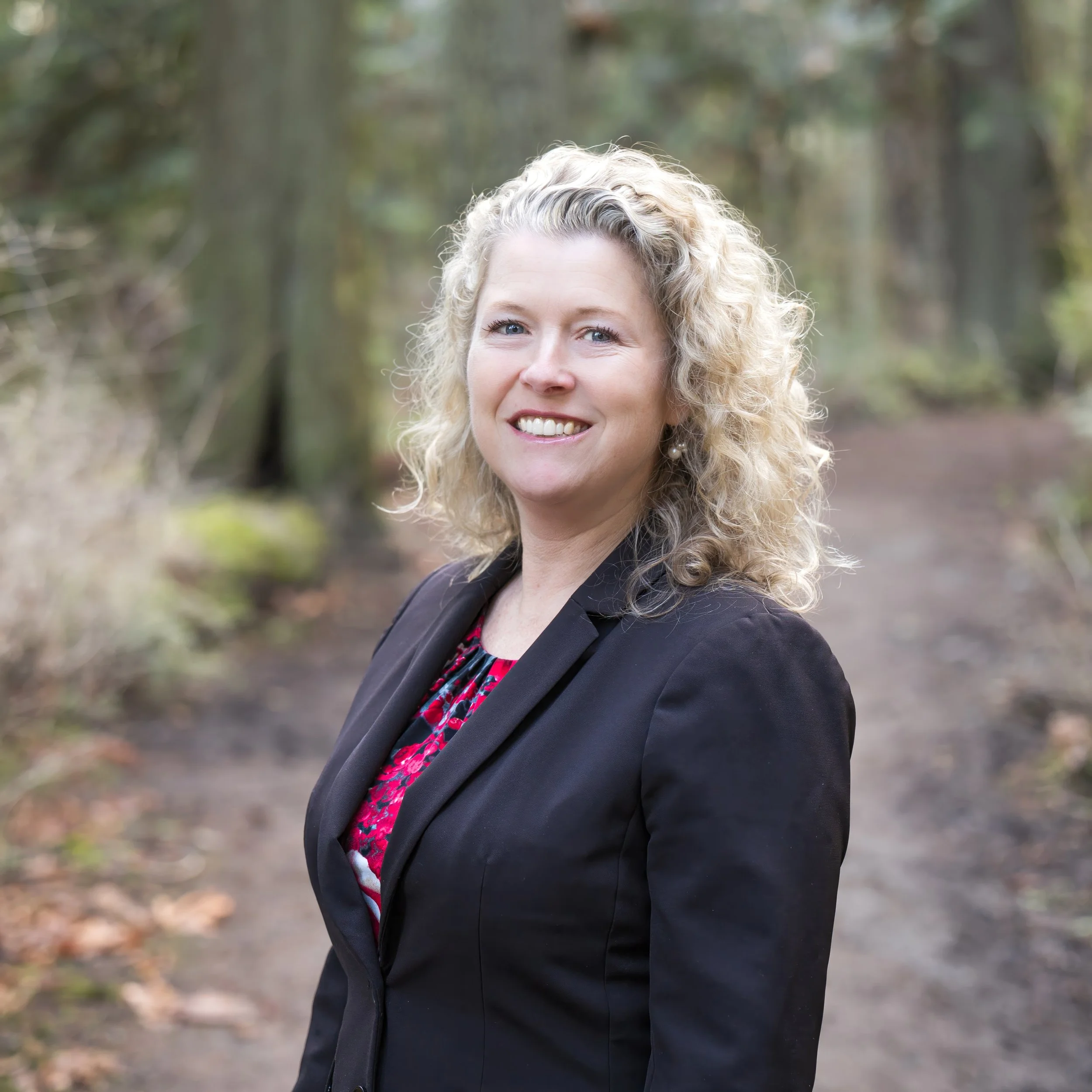Close-up of a woman with curly blonde hair, smiling, outdoors in a wooded area.