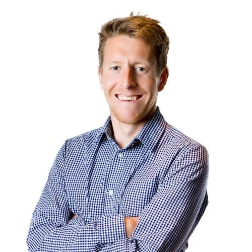 A smiling man with light skin and short ginger hair, wearing a blue and white checkered shirt, standing against a white background.