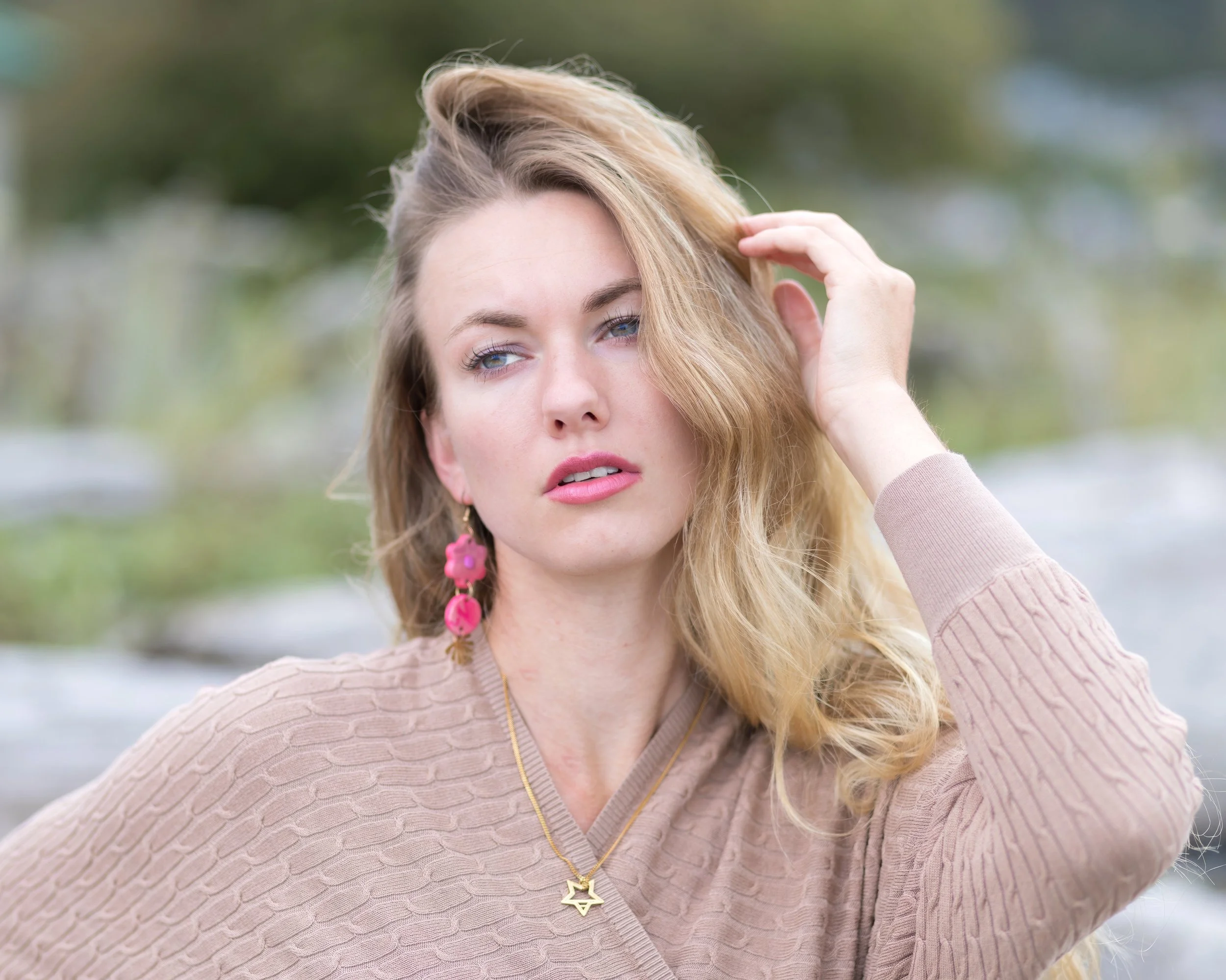 A woman with long blonde hair wearing pink earrings and layered necklaces, dressed in a textured beige sweater, poses outdoors with a blurred natural background.