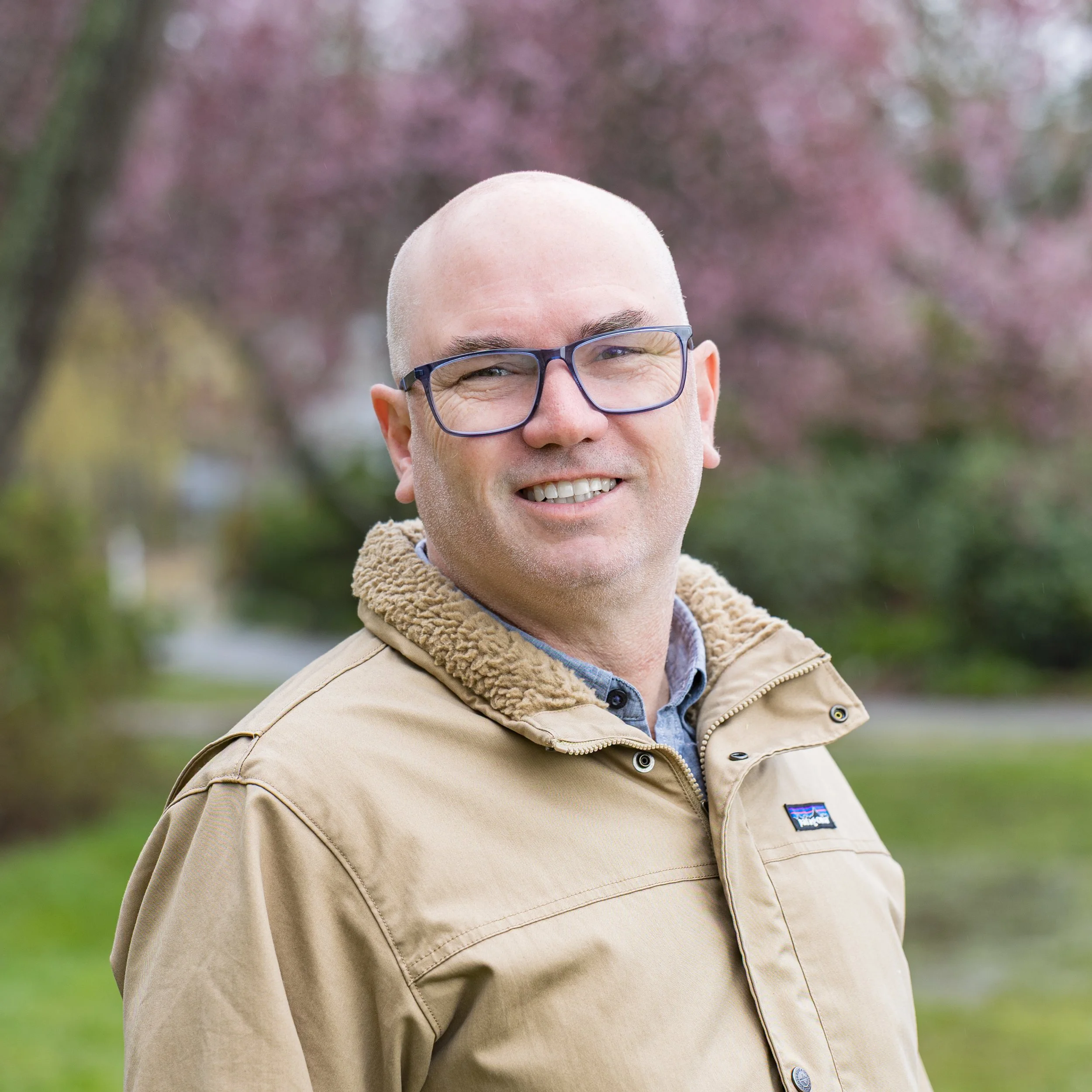 Headshot of a bald man wearing glasses and a gray button-up shirt, smiling with arms crossed against a plain white background.