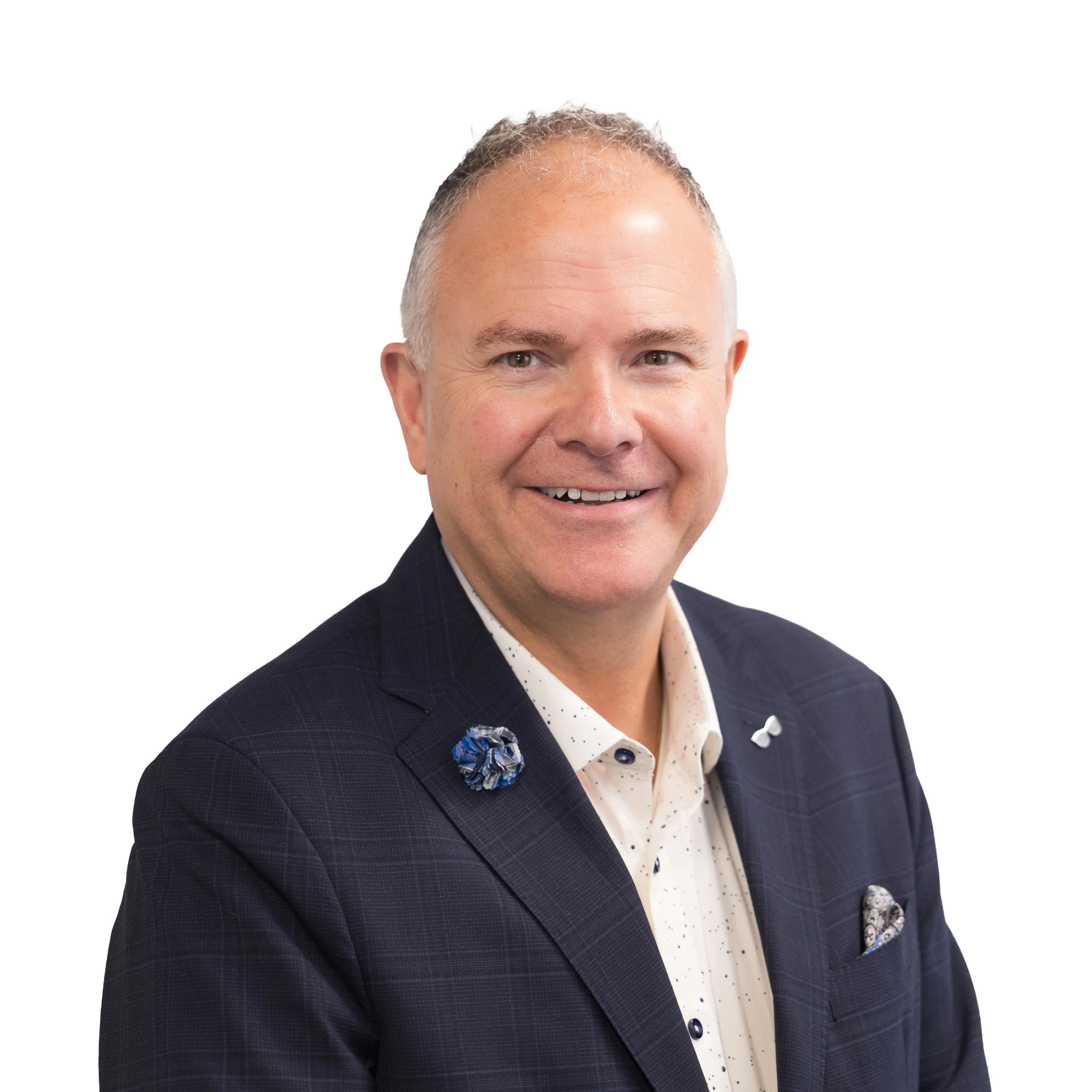 Close-up of a smiling mature man with short gray hair, wearing a dark checkered blazer, a white shirt with small dark patterns, and a blue patterned pocket square, set against a plain white background.