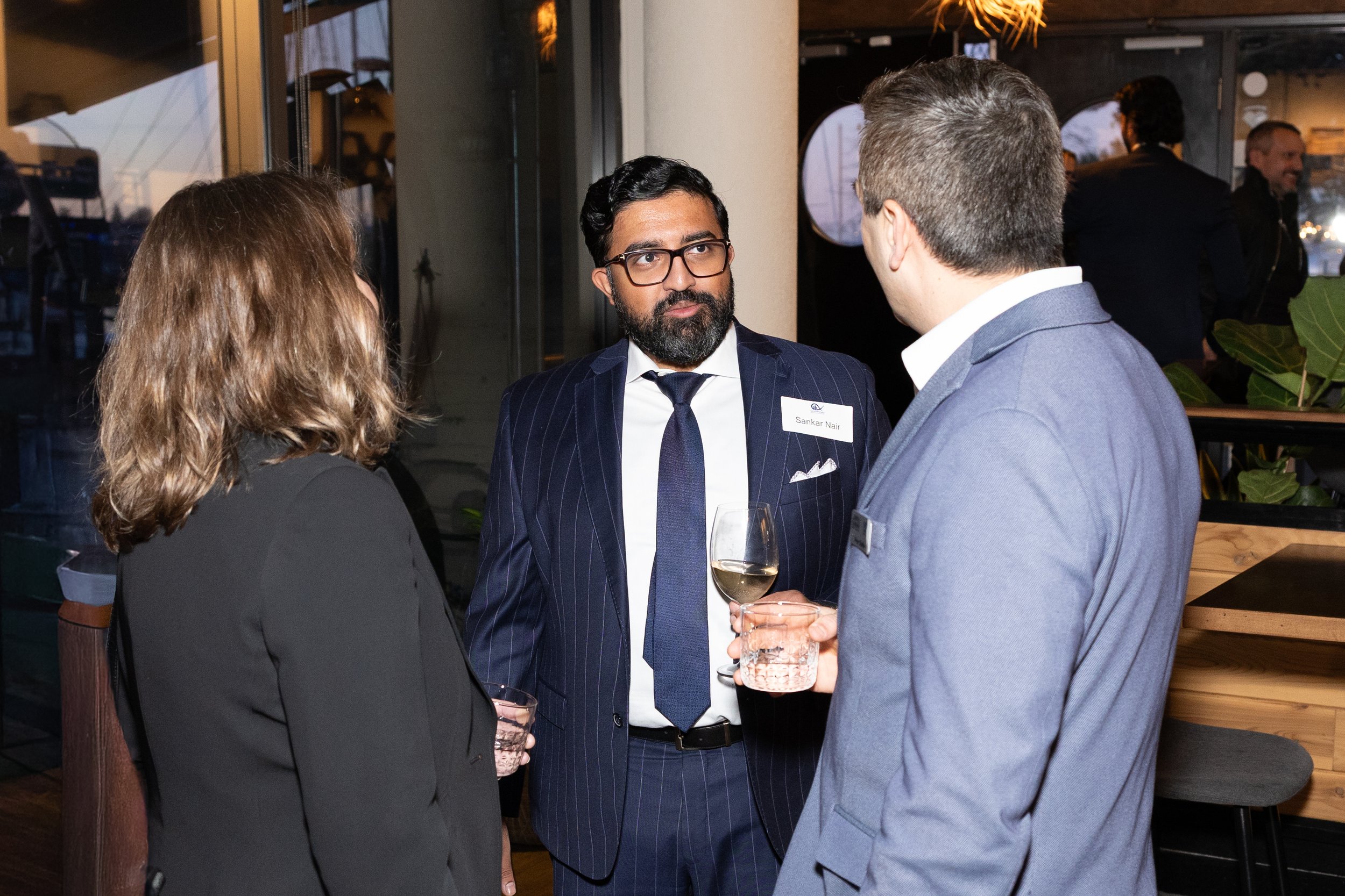Three people in business attire having a conversation at a social event indoors. One man with a beard and glasses is facing the camera, wearing a striped navy suit and a name tag. A woman with shoulder-length blonde hair and a man with short hair and