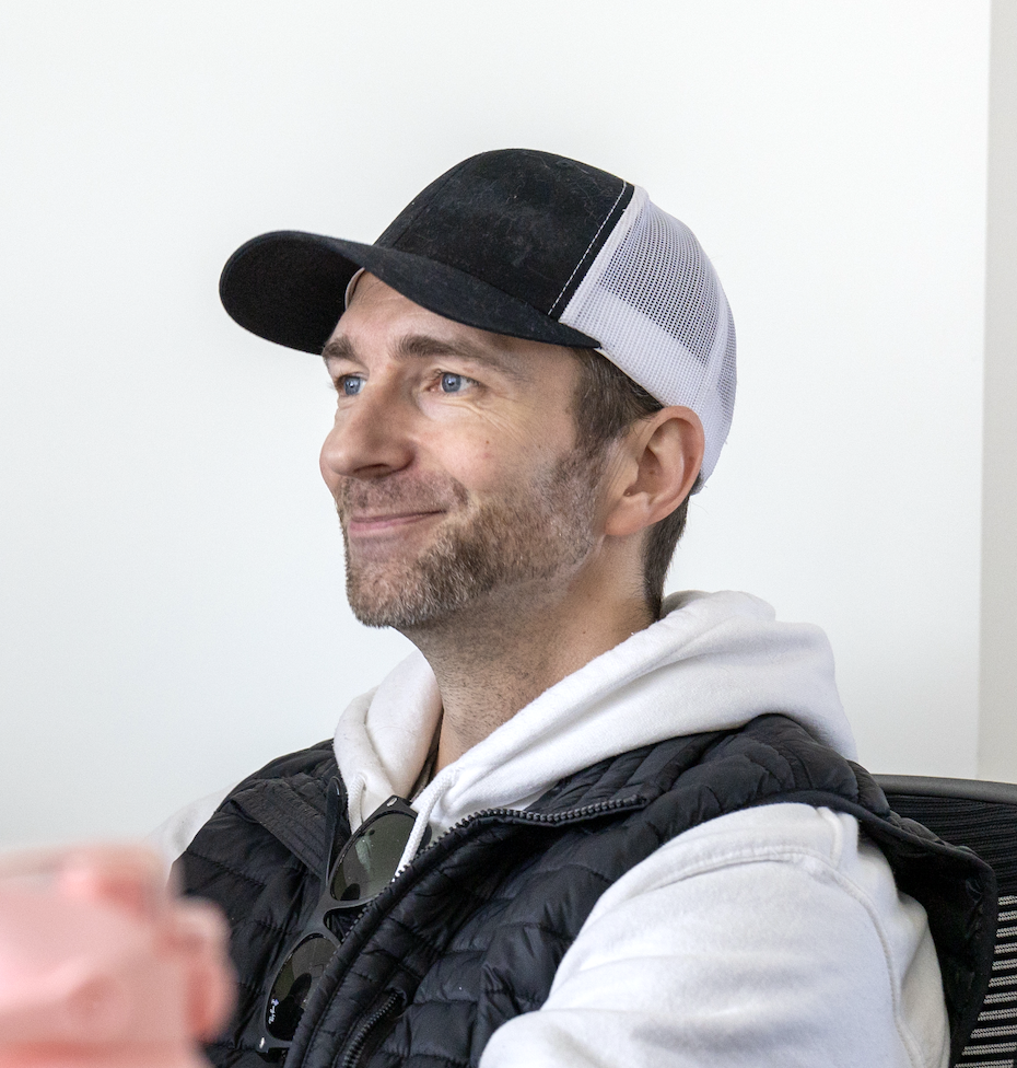 A man wearing a white hoodie, black vest, and a black and white baseball cap, smiling while looking to the side.