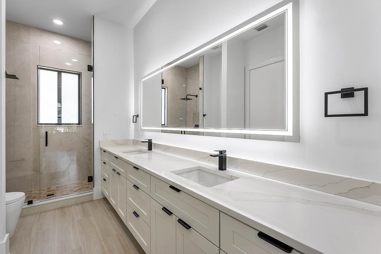 Modern white bathroom with double vanity and black fixtures, large backlit mirror, walk-in shower with glass door, and a window.