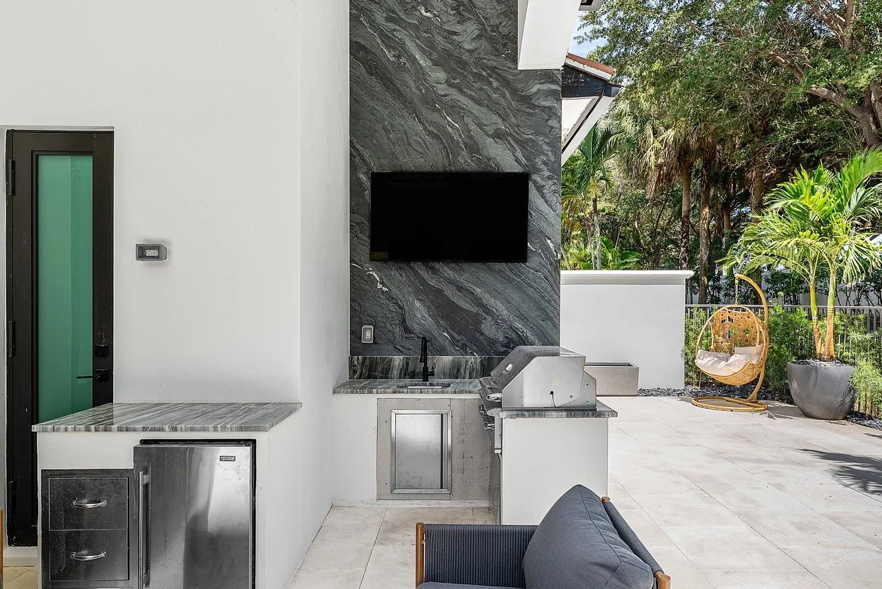 Outdoor patio area with a built-in grill, a mounted flat-screen TV, a small sink, and a stone countertop. There is a black door to the left, a chair in the foreground, and lush greenery with palm trees in the background.