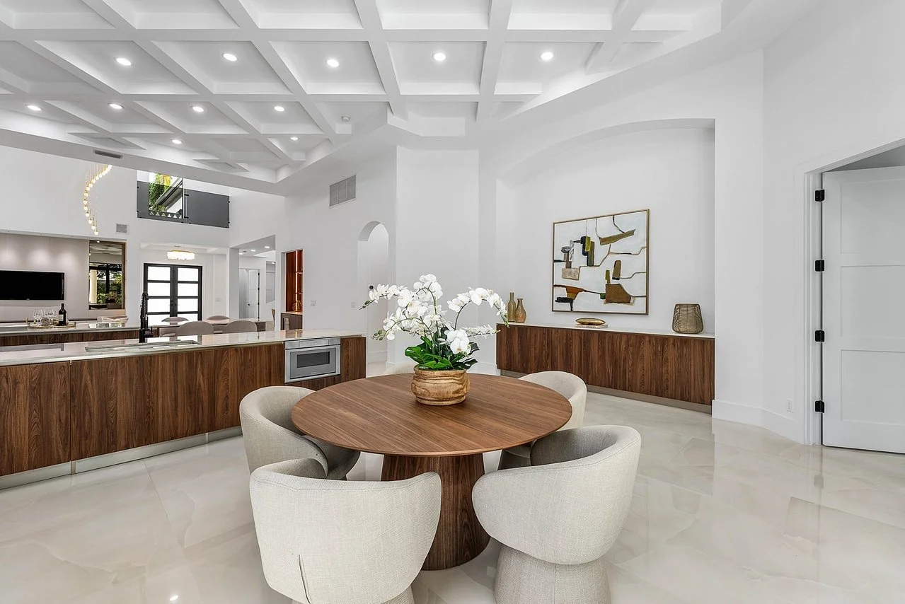 Modern dining area with a round wooden table, beige upholstered chairs, white orchid centerpiece, white walls, wooden sideboard, abstract wall art, and a coffered ceiling with recessed lighting.