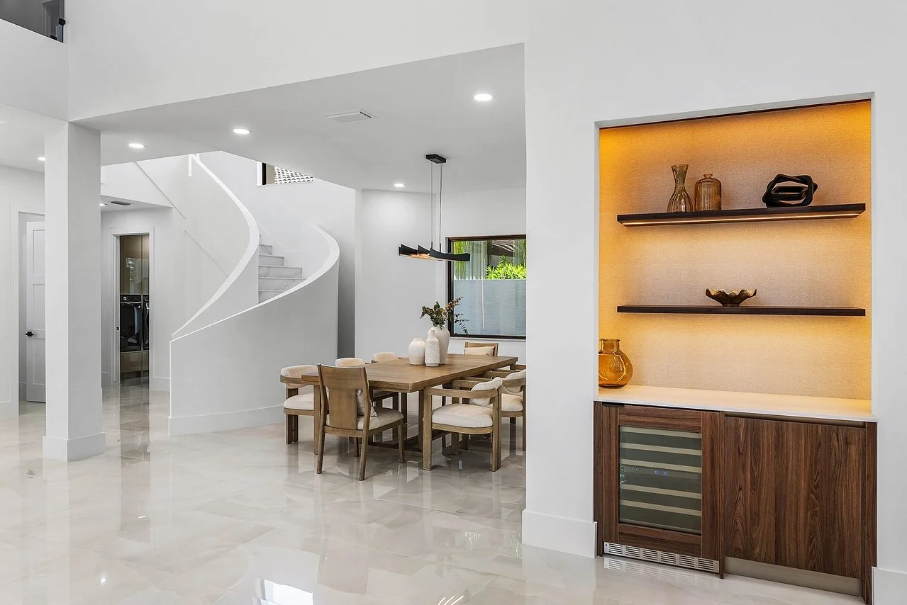 Modern dining area with a wooden table, beige chairs, and decorative vases, in a bright white interior with a spiral staircase in the background and built-in shelves with decorative items and a wine cooler.