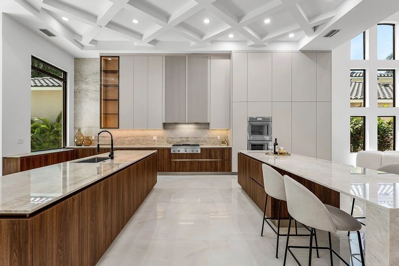 Modern kitchen with white cabinets, wooden accents, marble countertops, and a coffered ceiling, featuring a large island with seating and stainless steel appliances.