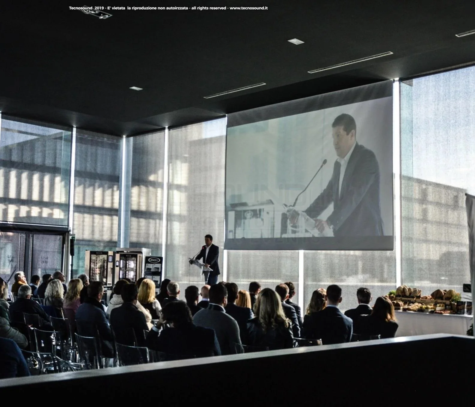 Un uomo in giacca e cravatta al microfono davanti a un pubblico durante una conferenza, con una grande proiezione di lui sullo sfondo all'interno di una sala moderna con grandi finestroni.