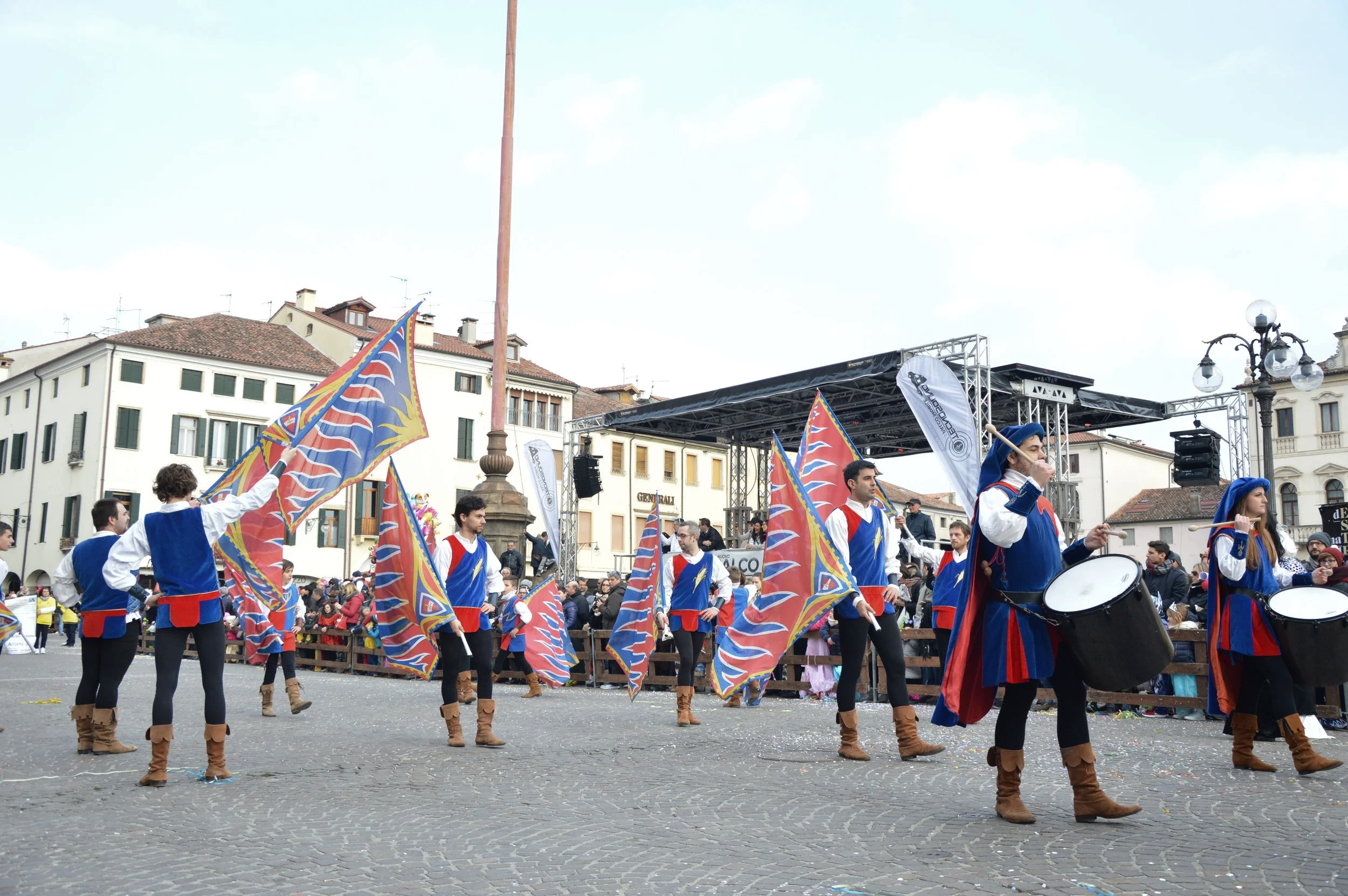 Gruppo di persone in costume medievale con bandiere colorate che sfilano in piazza durante una manifestazione, con pubblico seduto e uno stage sullo sfondo.