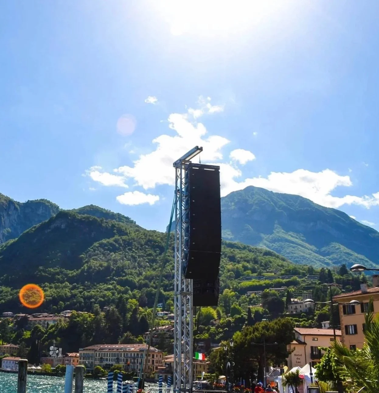 Montagna verde con case e, sullo sfondo, un grande schermo in una struttura metallica e un cielo azzurro con alcune nuvole.