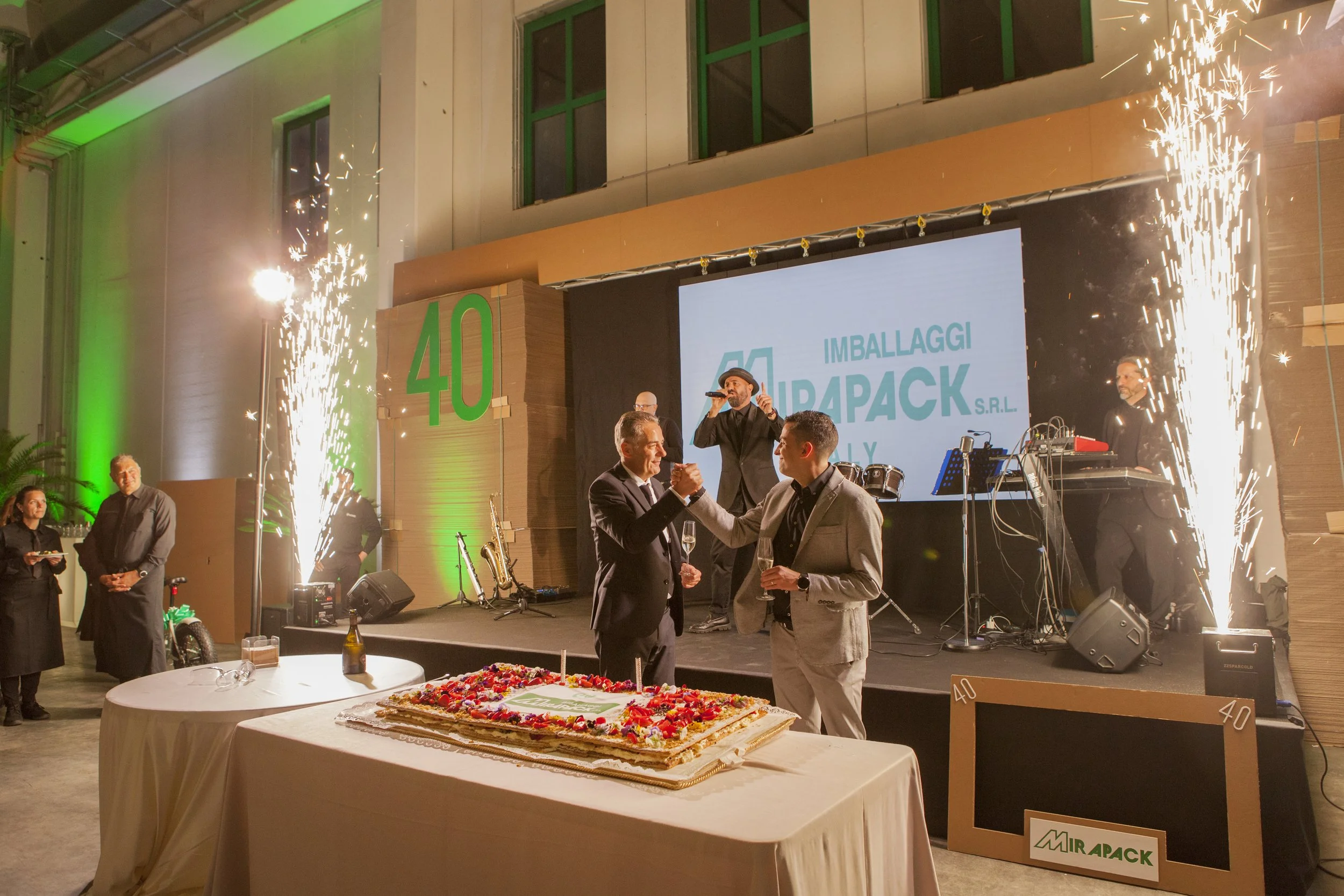 Celebrazione con torta e fuochi d'artificio in un evento aziendale, con due uomini che brindano e un gruppo musicale sul palco, decorazioni e grande numero 40.