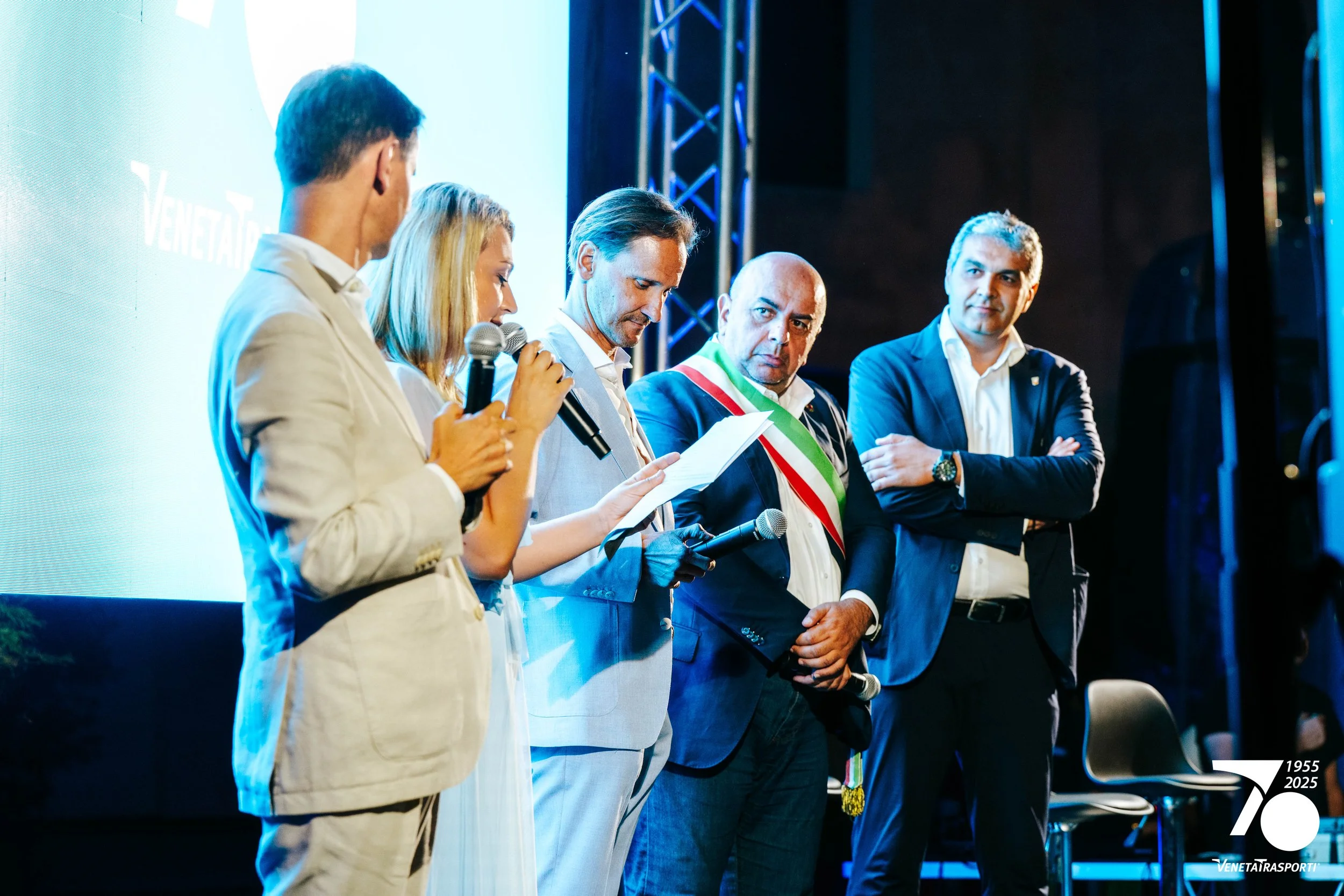 Un gruppo di cinque persone sul palco durante un evento, alcune con microfono, una con la fascia tricolore, sfondo blu e luci, logo 'Venetia Trasporti' e celebrazione del 70º anniversario.