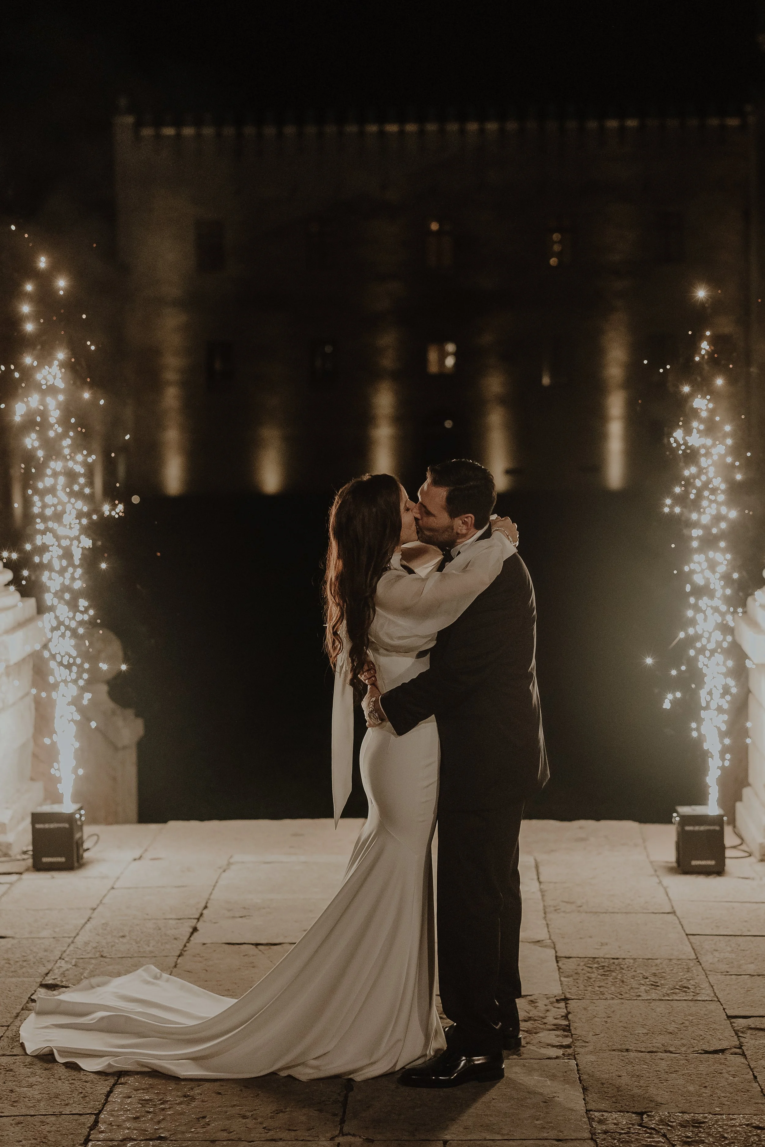 Una coppia di sposi si bacia romanticamente di sera con fuochi d'artificio alle spalle, davanti a un castello illuminato.