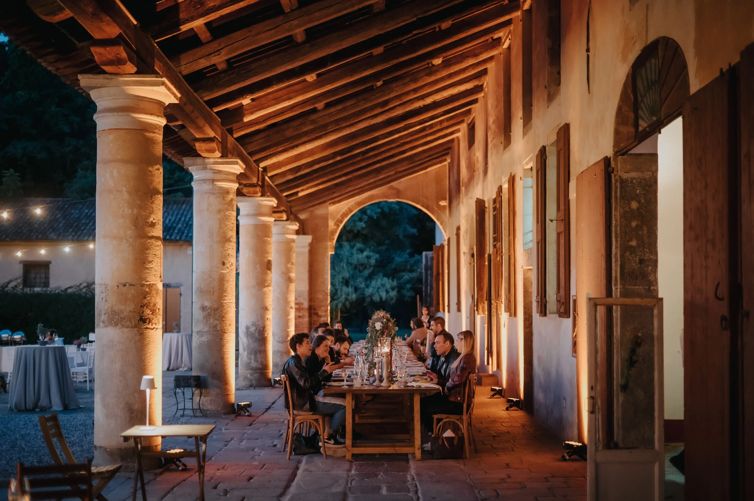 Cena all'aperto in un'atmosfera calda e rustica con persone sedute intorno a un lungo tavolo, decorato con fiori, sotto un porticato con colonne e luci soffuse, in un ambiente serale.