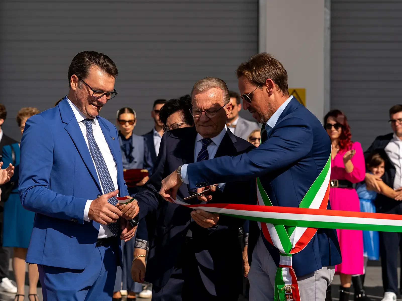 Uomo in abito blu che taglia un nastro tricolore durante una cerimonia ufficiale con altri partecipanti sorridenti e vestiti eleganti, in un'area industriale o di inaugurazione.