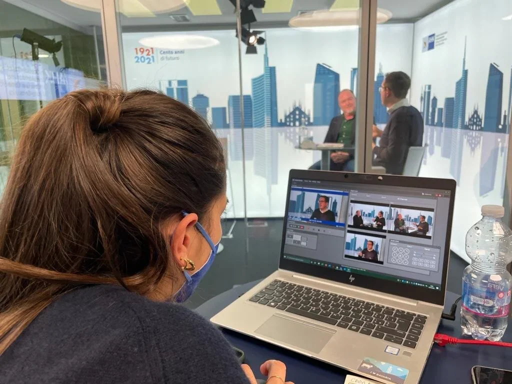 Una donna con mascherina sta registrando una intervista in uno studio con vista sulla città, con due uomini seduti a un tavolo dall'altra parte del vetro.