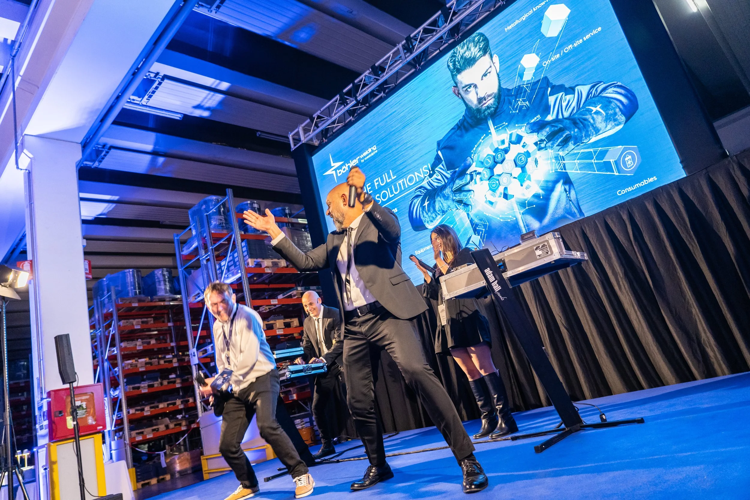 Persone che ballano e si divertono su un palco durante un evento, con uno schermo gigante sullo sfondo che mostra un uomo con una barba e una giacca con l'esposizione di tecnologie e soluzioni.