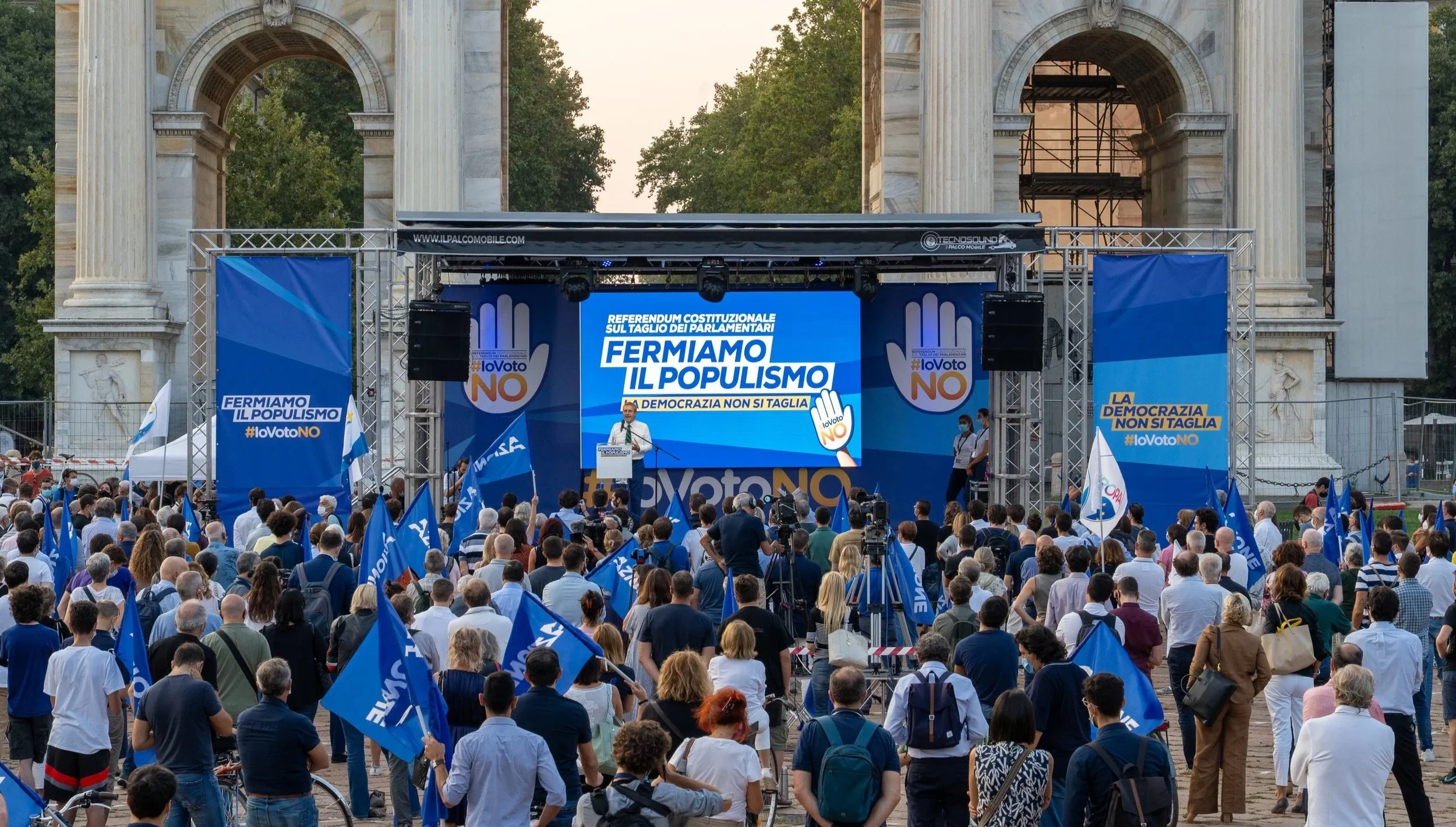 Manifestazione politica con molte persone che tengono bandiere blu con scritte 'Voto NO' e partecipano a un comizio di fronte a un monumento storico, con schermo e pannelli pubblicitari contro il populismo, in italiano.