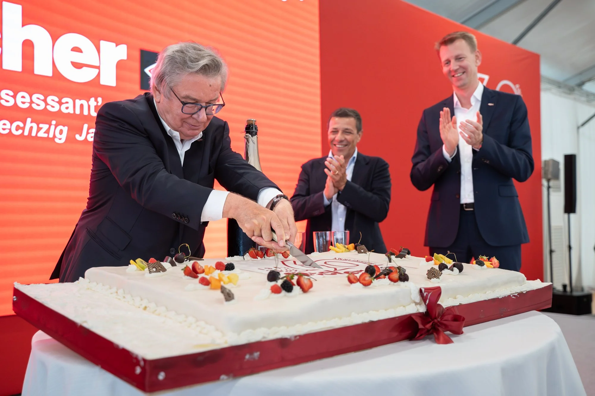 Un uomo anziano con capelli grigi e occhiali taglia una grande torta decorata con frutta e cioccolato, mentre due uomini più giovani applaudono e sorridono in un contesto celebrativo.