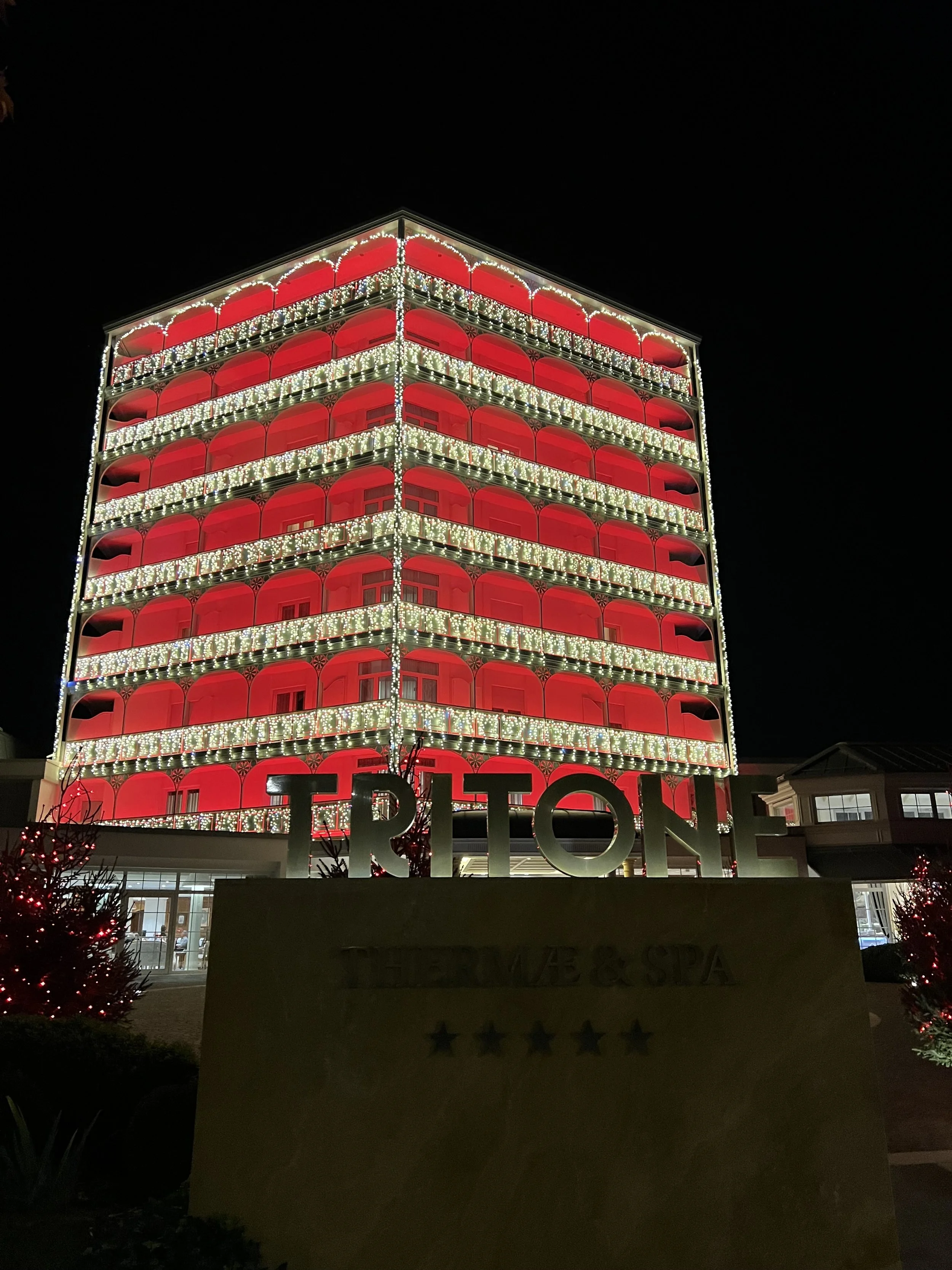 Edificio illuminato con luci natalizie rosse e bianche, decorato per le festività, con il nome 'TRITONE' in primo piano.