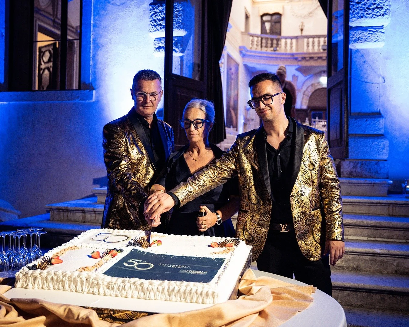 Tre persone, due uomini e una donna, tagliano una torta di compleanno durante una festa, indossando abiti eleganti con giacche dorate, all'aperto in una location elegante.