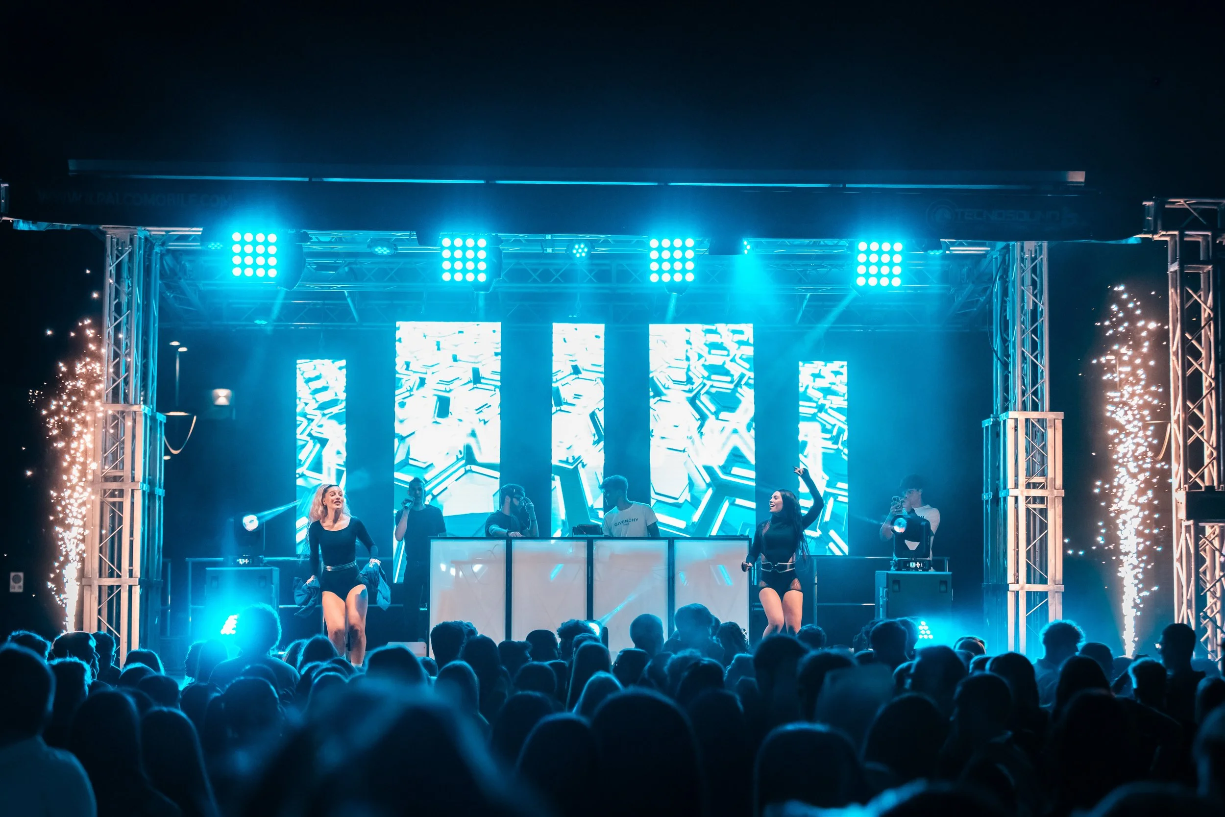 Performance di musica elettronica con due ballerini e DJ in scena, luci blue e effetti di scintille in un concerto all'aperto di notte.