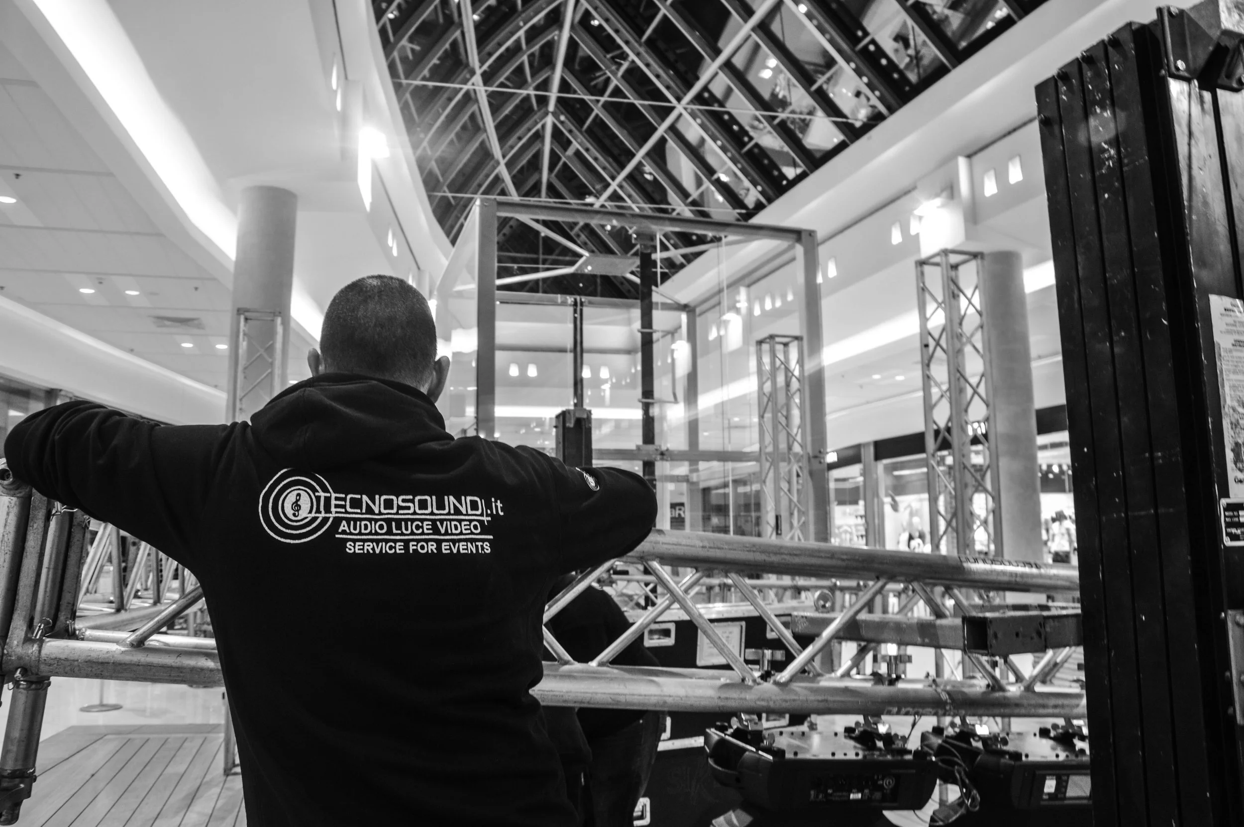 Una persona con felpa con logo Tecnosound, lavora su un impianto audio e luci in un centro commerciale, con strutture in metallo e vetro al soffitto.