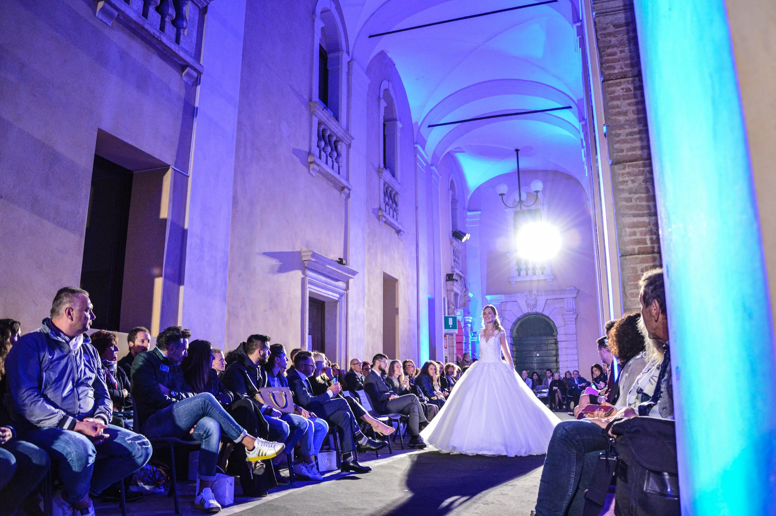Modella in abito da sposa sfilando davanti a un pubblico seduto, in una grande sala con archi e pareti illuminate di blu.