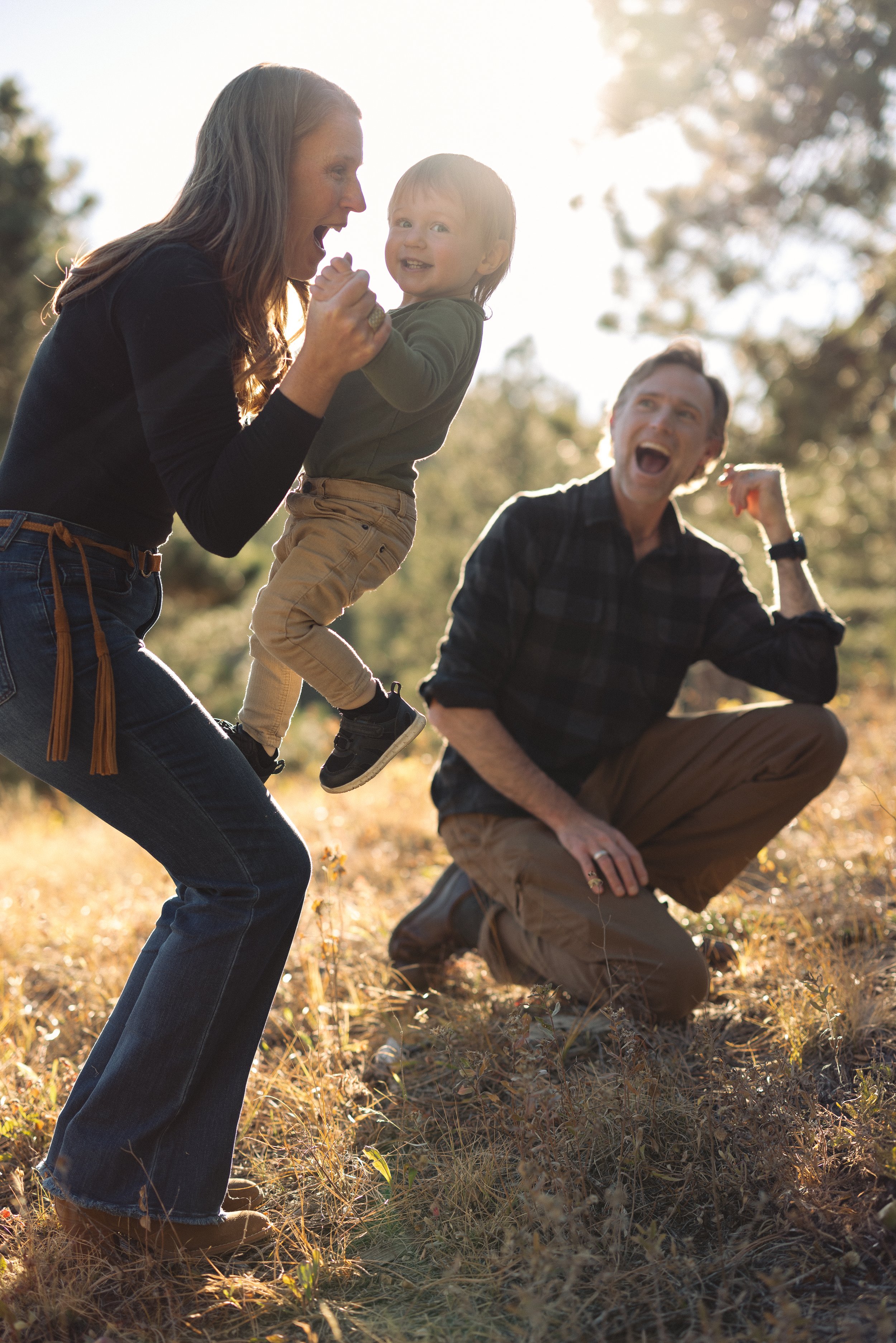 A family of three having fun outdoors in a park during autumn, with the mother lifting her young son, and the father kneeling on one knee, all smiling and enjoying the moment.